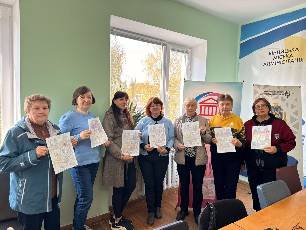 Переселенки з Херсонщини тримають у руках свої патріотичні малюнки, позуючи в кабінеті вінницького координаційного центру «Вільні разом» під час антистресового малювання «Запорізька Січ», яке провели в межах Тижня ментального здоров’я. Жінки старшого та середнього віку вишикувалися в один ряд біля великого вікна та посміхаються в об'єктив кадру, демонструючи деталізовані розмальовки із зображенням українських козаків на тлі старовинної фортеці. Учасниці зустрічі, вбрані в повсякденні осінні куртки, светри та кофти, тримають свої готові творчі роботи перед собою для спільної пам'ятної світлини. Заходи з психоемоційного розвантаження, які також включали психологічну гру «Мої трансформації» та ресурсну зустріч «Випадковість, яка веде», проходять у світлій кімнаті хабу на тлі великого настінного банера з написом «ВІННИЦЬКА МІСЬКА АДМІНІСТРАЦІЯ» та прапора «ХЕРСОНСЬКА ОБЛАСНА ДЕРЖАВНА АДМІНІСТРАЦІЯ» із золотим тризубом і офіційним гербом області праворуч.