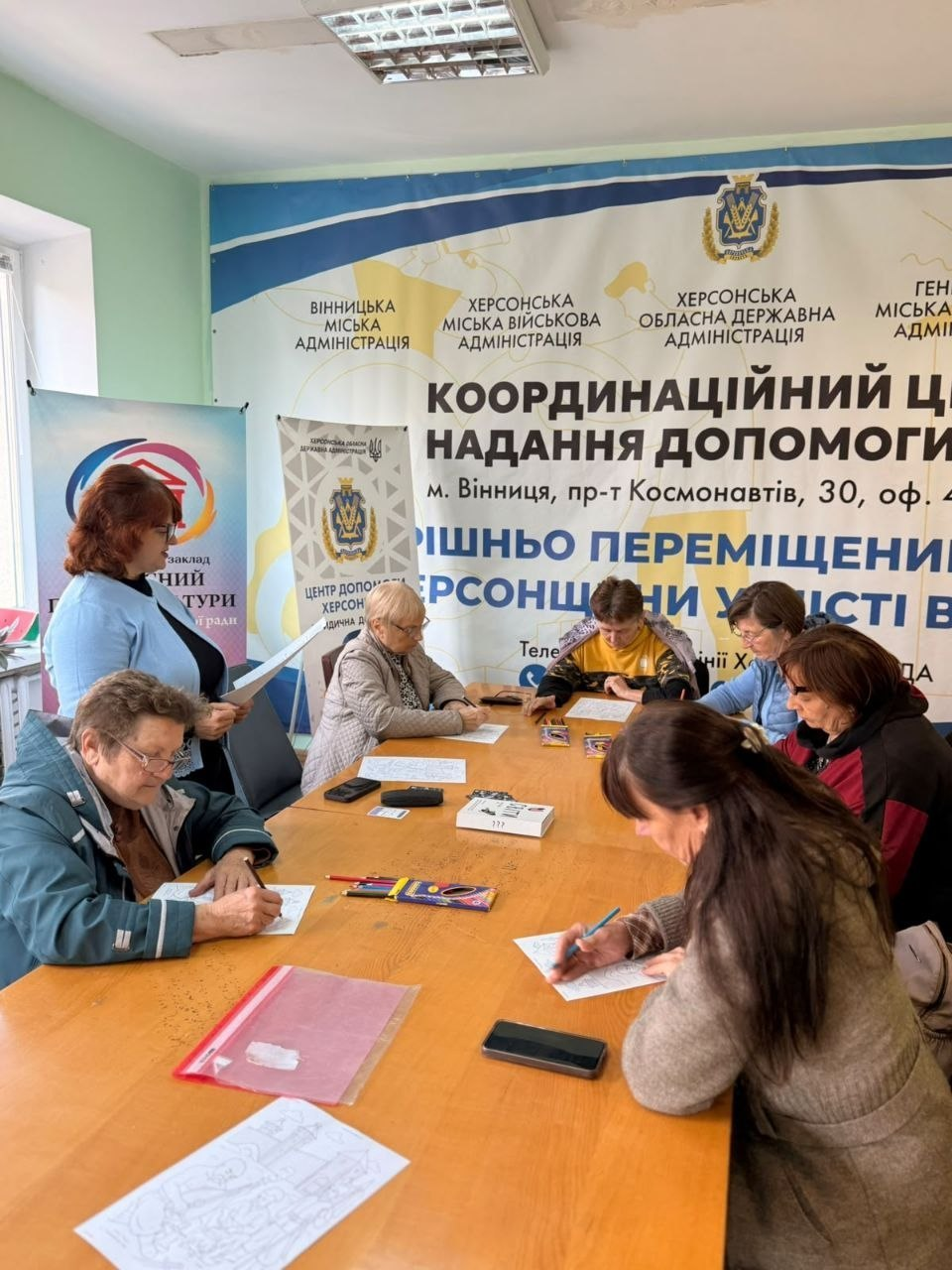Переселенки з Херсонщини зосереджено розмальовують патріотичні малюнки, сидячи за великим спільним столом у вінницькому координаційному центрі «Вільні разом» під час антистресового заняття «Запорізька Січ», організованого в межах Тижня ментального здоров’я. Жінки різного віку в кофтах та куртках роблять нотатки й охайно розфарбовують ескізи козацької тематики кольоровими олівцями та маркерами, розкладеними посеред довгого дерев'яного столу поруч із мобільними телефонами та бланками. Ліворуч біля вікна стоїть рудоволоса координаторка в блакитній кофті, яка переглядає робочі документи та спілкується з присутніми для їхнього психоемоційного розвантаження й емоційної підтримки в евакуації. Комплексна ментальна програма, яка також включала психологічну гру «Мої трансформації» та ресурсну зустріч «Випадковість, яка веде», проходить у світлому кабінеті хабу на тлі великого офіційного банера з гербом і текстом «КООРДИНАЦІЙНИЙ ЦЕНТР НАДАННЯ ДОПОМОГИ ВНУТРІШНЬО ПЕРЕМІЩЕНИМ ХЕРСОНЦЯМ У МІСТІ ВІННИЦЯ», а також прапорів Вінницької, Херсонської, Генічеської міських та обласних військових адміністрацій.