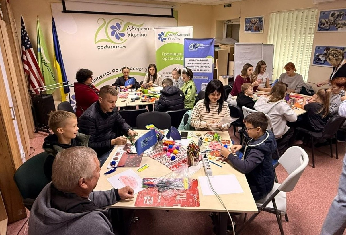 Дорослі та діти з родин переселенців із Херсонщини захоплено створюють яскраві саморобки у вінницькому координаційному центрі «Вільні разом» під час сімейного майстер-класу «Мої пухнасті фантазії». У залі хабу учасники розмістилися групами навколо трьох столів, де разом із координаторами скручують фігурки з м'якого синельного дроту для психоемоційного розвантаження сімей ВПО. На столах розкладено пухнасті помпони, кольорові олівці, клей та канцелярське приладдя для рукоділля. Цей арт-терапевтичний захід проходить у партнерстві з ГО «Джерело надії України», чиї фірмові білі та сині банери з гаслом «Розвиваємо. Впливаємо. Змінюємо(ся)» прикрашають стіну кімнати, де ліворуч біля вікна також встановлено Державні прапори України та Сполучених Штатів Америки.
