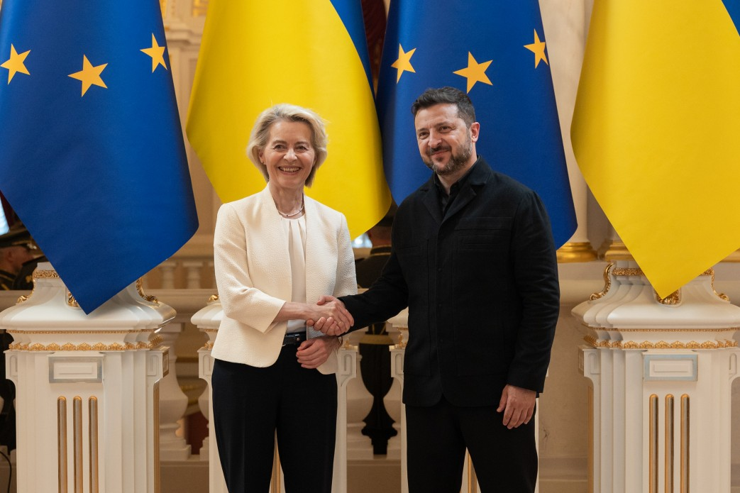 Президент України Володимир Зеленський та Президентка Європейської комісії Урсула фон дер Ляєн позують для протокольного фото під час рукостискання на тлі Державних прапорів України та стягів Євросоюзу. Урсула фон дер Ляєн у білому жакеті й темних брюках та Володимир Зеленський у чорному френчі уклали стратегічне партнерство, підписавши Лист про наміри щодо Drone Deal. Цей крок посилив євроінтеграційний рух України після відкриття переговорних кластерів про вступ та ухвалення 20 пакетів санкцій проти РФ. З початку великої війни ЄС став найбільшим донором нашої держави, надавши понад 216 мільярдів євро підтримки, включаючи пакет на 90 мільярдів євро на 2026–2027 роки, тренування воїнів, залучення надприбутків від заморожених активів Росії для ОПК та розбудову Спеціального трибуналу проти агресора.