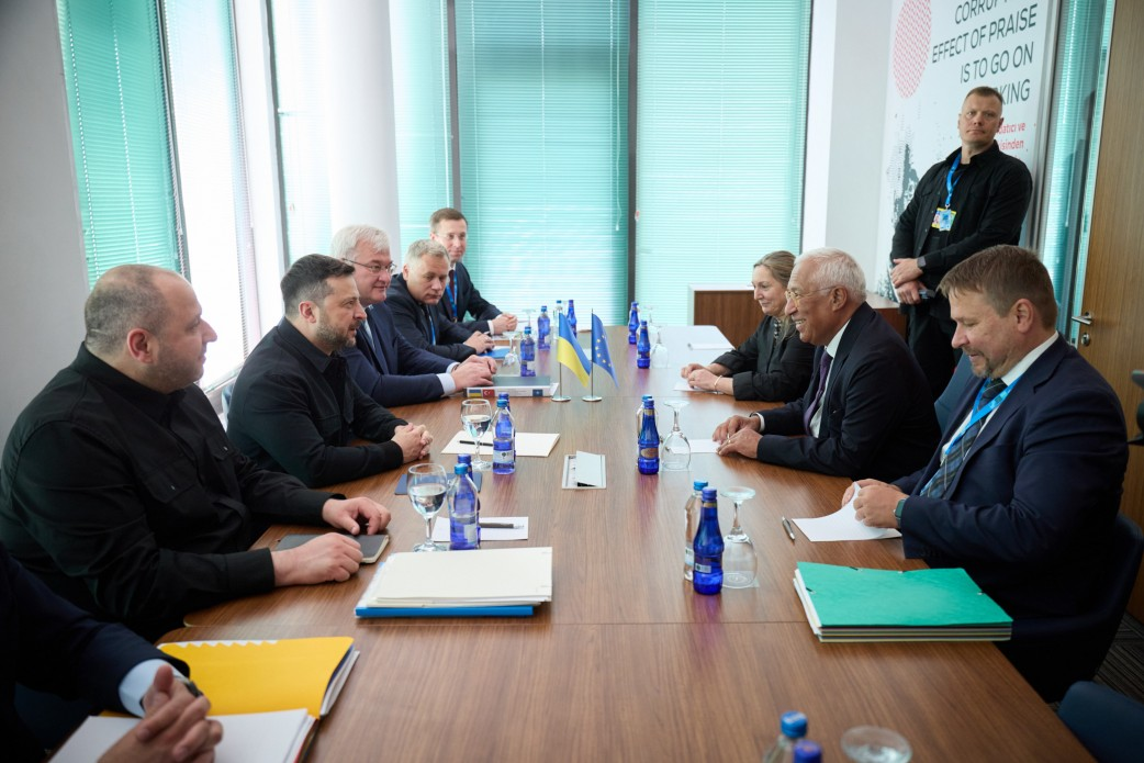 Президент України Володимир Зеленський та Президент Європейської ради Антоніу Кошта разом із делегаціями проводять переговори за столом в Анкарі, де в кадрі видно верхню частину тіла присутніх. Ліворуч сидить українська сторона на чолі з Володимиром Зеленським у чорному френчі та Міністром оборони Рустемом Умєровим, а на протилежному боці — Антоніу Кошта у темному костюмі з європейськими представниками. Посередині столу встановлено настільні прапори України та ЄС, розставлено пляшки з водою й документи, а на задньому плані за скляними дверима помітно чоловіка біля великого білого плаката. Під час зустрічі Глава держави подякував Антоніу Кошті за підтримку нашої країни на шляху до ЄС, а лідери обговорили якомога швидше відкриття решти п’яти переговорних кластерів і потенційний графік наступних кроків, на які заслуговує український народ.