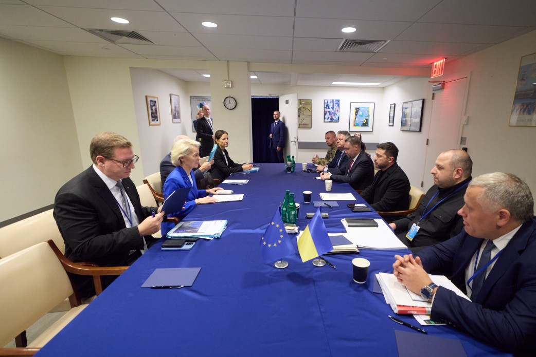 Президент України Володимир Зеленський та Президентка Європейської комісії Урсула фон дер Ляєн разом із членами офіційних делегацій проводять переговори за довгим столом під синьою скатертиною, де в кадрі видно верхню частину тіла присутніх. Праворуч сидить українська сторона на чолі з Володимиром Зеленським у чорному френчі, Міністром оборони Рустемом Умєровим та іншими посадовцями, а на протилежному боці — Урсула фон дер Ляєн у яскраво-блакитному жакеті з європейськими представниками. Посередині столу встановлено настільні прапори Євросоюзу та України, розставлено пляшки з водою й папки з документами, а на задньому плані кімнати помітно відкриті білі двері та розвішані на стінах картини й фотографії. Під час цієї зустрічі лідери детально обговорили, як забезпечити фінансову стійкість України цього та наступного року, при цьому Глава держави подякував за рішення щодо позики ЄС у 90 мільярдів євро для максимального закриття бюджетного дефіциту. Президенти домовилися про опрацювання командами інших шляхів фінансової підтримки, а також зосередили особливий фокус на санкціях Євросоюзу проти Росії, де Володимир Зеленський наголосив на самовбивчості та контрпродуктивності сигналів про зняття обмежень з окремих російських олігархів і подякував Урсулі фон дер Ляєн за готовність попрацювати над включенням додаткових прізвищ наближених агресора до санкційних списків.