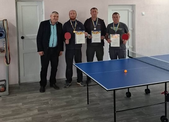 У Борозенській громаді відбулись змагання з настільного тенісу, які присвятили Дню захисників та захисниць України. На пам'ятній світлині з турніру на Бериславщині чотири чоловіки позують для спільного фото на повний зріст у спортивному залі за синім тенісним столом із помаранчевою кулькою та натягнутою сіткою. Ліворуч стоїть організатор заходу, який також є представником громади, у класичному чорному піджаку та блакитній сорочці, а поруч із ним вишикувалися троє переможців турніру у спортивному одязі з медалями на шиї, які тримають у руках почесні грамоти та червоні ракетки для пінг-понгу на тлі рожевих стін та білих дверей залу. У поєдинках за тенісними столами зійшлись досвідчені гравці та початківці, кожен із яких продемонстрував належну майстерність і жагу до перемоги, а за результатами запеклих партій кращі учасники отримали заслужені нагороди та дипломи від громади.