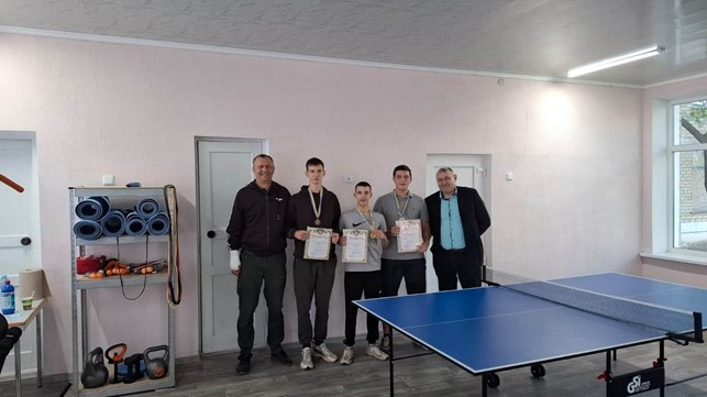 Двоє організаторів турніру та троє юнаків-переможців змагань із настільного тенісу позують для спільного пам'ятного фото на повний зріст у спортивному залі Борозенської громади, вишикувавшись в один ряд за великим синім тенісним столом марки GSI-Sport із натягнутою сіткою. З лівого краю кадру стоїть перший організатор заходу у темній спортивній кофті з перев'язаною білим бинтом лівою рукою та в зелених штанях, а з правого краю ряд замикає другий організатор, який також є представником громади, у класичному чорному піджаку та блакитній сорочці. По центру між ними вишикувалися три юні призери у спортивних костюмах, футболках та худі з медалями на шиї, які тримають у руках отримані нагородні дипломи та почесні грамоти. Спільний знімок зроблено у затишному світлому приміщенні з сірим ламінатом на підлозі, рожевими стінами, двома зачиненими білими дверима на задньому плані, великим вікном праворуч та металевим стелажем ліворуч, де складено сині фітнес-килимки й гирі.