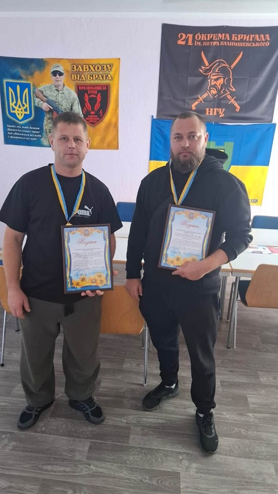Двоє учасників змагань із настільного тенісу позують для спільного фото на повний зріст у приміщенні з сірим ламінатом на підлозі під час турніру в Борозенській громаді. Ліворуч стоїть чоловік у чорній футболці з білим логотипом Puma та широких зелених штанях, а праворуч від нього стоїть чоловік із густою бородою, вбраний у чорне худі та темні спортивні штані. Обидва призери тримають перед собою нагородні грамоти у тонких темних рамках, а на їхніх шиях зафіксовано медалі на синьо-жовтих стрічках. Патріотичний знімок зроблено на тлі світло-рожевої стіни, де позаду чоловіків розвішано синьо-жовтий Державний прапор України із зеленим щитом громади по центру, чорний військовий прапор із помаранчевим логотипом воїна та великим написом «21 окрема бригада ім. Петра Калнишевського НГУ», а також жовтий подарунковий банер із зображенням військового у сонцезахисних окулярах та написами «Завхозу від брата» і «Віра сильніша за страх».