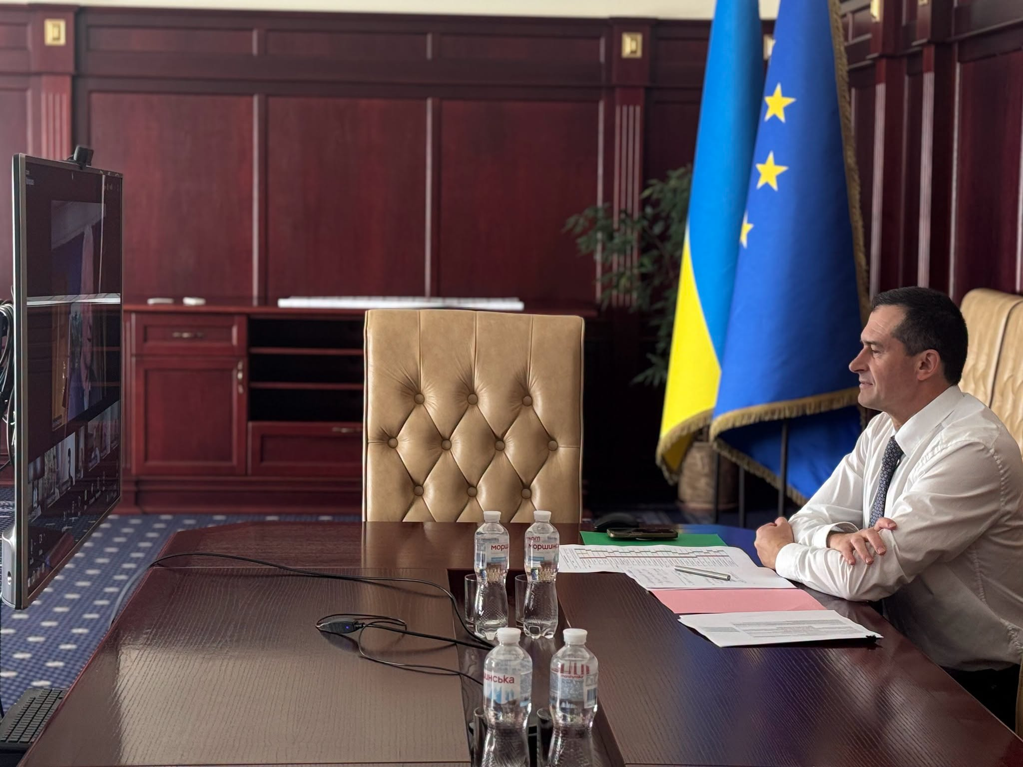 Віцепрем’єр-міністр Всеволод Ченцов у білій сорочці та краватці сидить у профіль за великим дерев'яним столом під час онлайн-зустрічі з Генеральним директором Генерального директорату Єврокомісії з питань розширення Гертом Яном Купманом. Посадовець склав руки перед собою й дивиться на вертикальний монітор відеозв'язку ліворуч, на столі розставлено пляшки води «Моршинська» й лежать документи поруч із порожнім бежевим кріслом, а за його спиною на тлі дерев'яних панелей кабінету височіють прапори України та ЄС. Під час перемовин сторони обговорили кроки для прискорення вступу України до Євросоюзу, приділивши увагу роботі над проміжними критеріями в межах Кластера 1 та підготовці до звіту IBAR. Всеволод Ченцов наголосив на важливості належної координації між Урядом і Парламентом, а також сторони домовилися просувати рішення для інтеграції у платіжний простір SEPA, внутрішній енергетичний ринок ЄС та використання можливостей штучного інтелекту для адаптації національного законодавства.