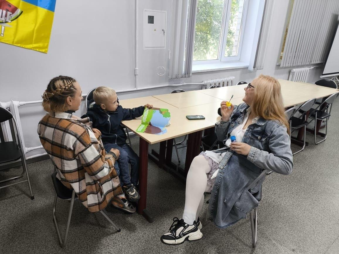 Логопединя-дефектологиня проводить індивідуальне розвивальне заняття для дитини в житомирському координаційному центрі «Вільні разом», який системно підтримує переселенців з Херсонщини. У світлому кабінеті за великим дерев'яним столом сидить хлопчик, який гортає яскраву дитячу книжку, поруч із мамою у кардигані в клітинку. Навпроти них із посмішкою сидить фахівчиня в окулярах та джинсовому плащі, яка пускає мильні бульбашки для емоційного розвантаження та розвитку артикуляції малюка, а на задньому плані помітно довгий ряд столів біля пластикового вікна. Під час цієї ініціативи спеціалістка провела корисні індивідуальні заняття для дорослих і дітей із родин внутрішньо переміщених осіб, допомагаючи херсонцям покращити мовленнєві навички, подолати стрес від евакуації та отримати необхідну підтримку.