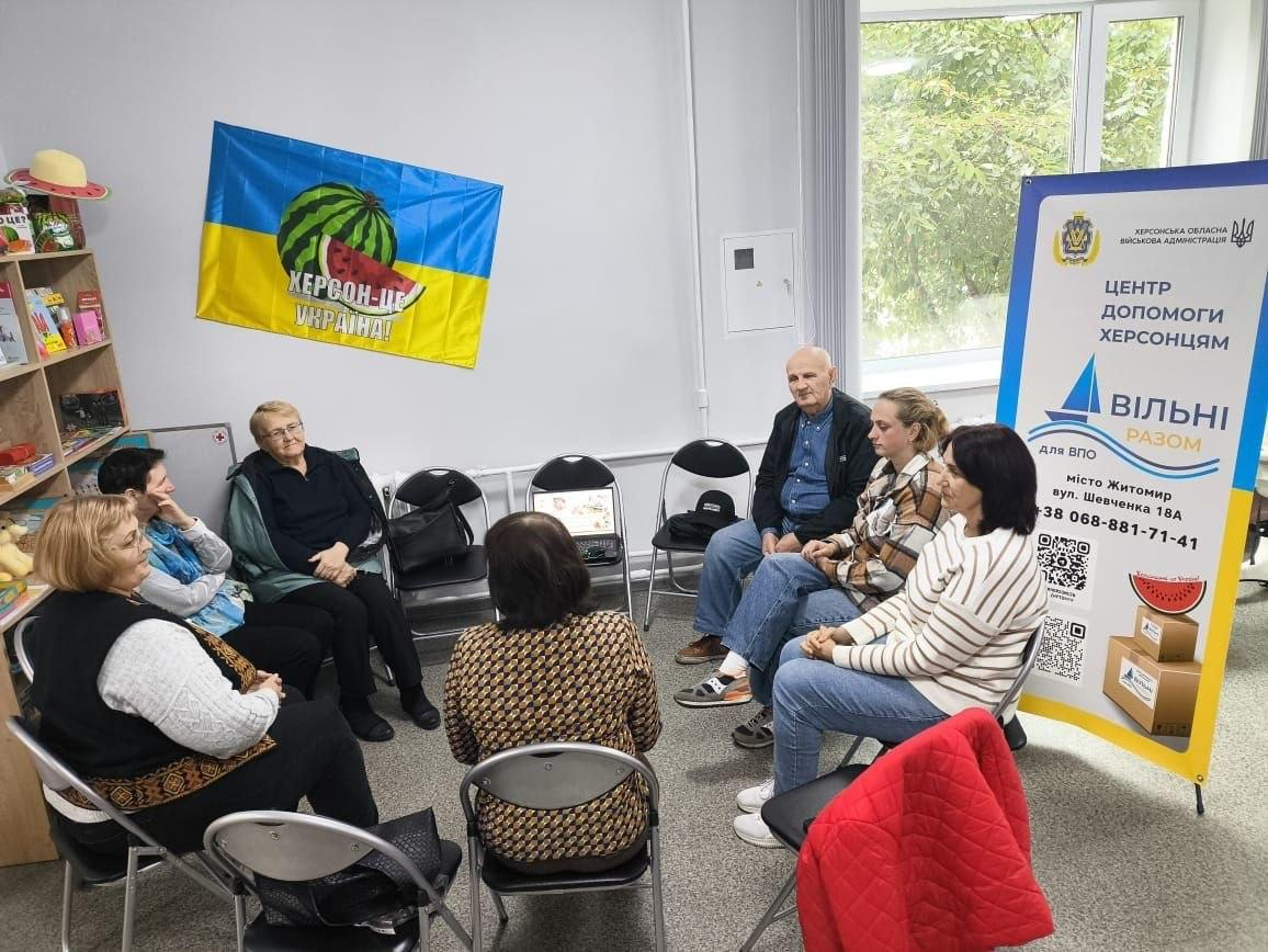 Координаційний центр «Вільні разом» у Житомирі провів для переселенців із Херсонщини заняття розмовного клубу «Українська - мова вільних, мова єднання» з Надією Гнатовською на тему фольклору та сучасних мемів. У кабінеті хабу учасники різного віку розсілися колом на стільцях для невимушеного спілкування, де ліворуч помітно полиці з книжками, а на задньому плані сидить літній чоловік біля ноутбука. На стіні над присутніми розгорнуто синьо-жовтий прапор із малюнком кавуна та написом «ХЕРСОН-ЦЕ УКРАЇНА!», а праворуч біля великого вікна встановлено фірмовий інформаційний банер ХОВА з контактами житомирського центру допомоги ВПО. Такі зустрічі допомагають землякам підтримувати психоемоційний стан в евакуації, удосконалювати мовні навички та зберігати тісний зв'язок із рідною культурою.