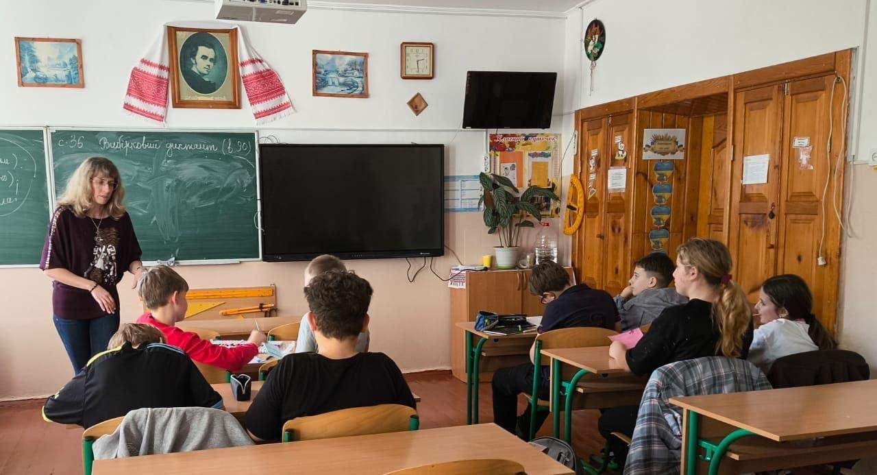 Координаційний центр «Вільні разом» у Житомирі організував заняття з психосоціальної підтримки для учнів 6 класу ліцею № 33, де разом із місцевими дітьми навчаються юні переселенці з Херсонщини. На світлині з кабінету української мови кучерява вчителька у темній блузці та джинсах стоїть біля зеленої дошки та пояснює матеріал школярам, які сидять за партами. У кабінеті встановлено великий вимкнений інтерактивний екран, стіну прикрашає портрет Тараса Шевченка з вишиваним рушником, а правий бік кімнати облаштовано дерев'яними шафами з кімнатною квіткою. Такі інтеграційні заходи за сприяння хабу допомагають дітям-переселенцям швидше адаптуватися до нового середовища, покращити емоційний стан в евакуації та знайти друзів серед однокласників.