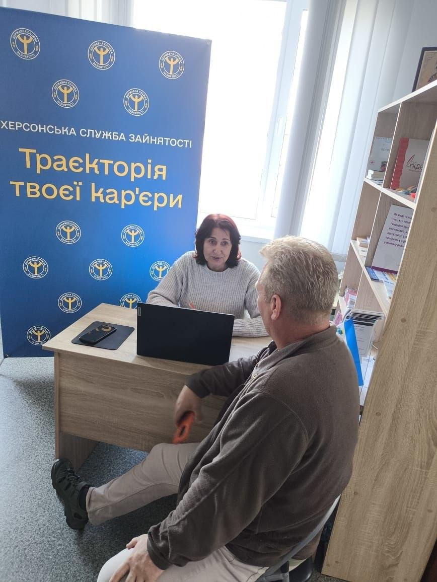 Фахівець Бериславської філії Херсонського обласного центру зайнятості проводить консультацію з питань працевлаштування для відвідувача в житомирському координаційному центрі «Вільні разом», який системно підтримує переселенців із Херсонщини. У світлому кабінеті за дерев'яним столом сидітиме жінка-консультант у світлому светрі з відкритим ноутбуком, яка спілкується з чоловіком у коричневій кофті. Ліворуч у кімнаті встановлено великий синій банер із написами «ХЕРСОНСЬКА СЛУЖБА ЗАЙНЯТОСТІ. Траєкторія твоєї кар'єри», а праворуч помітно високу дерев'яну шафу-стелаж з інформаційними буклетами. Під час цієї правової та професійної зустрічі внутрішньо переміщені особи отримали індивідуальні роз'яснення щодо актуальних вакансій, можливостей перенавчання та отримання грантів для успішного пошуку роботи в евакуації.