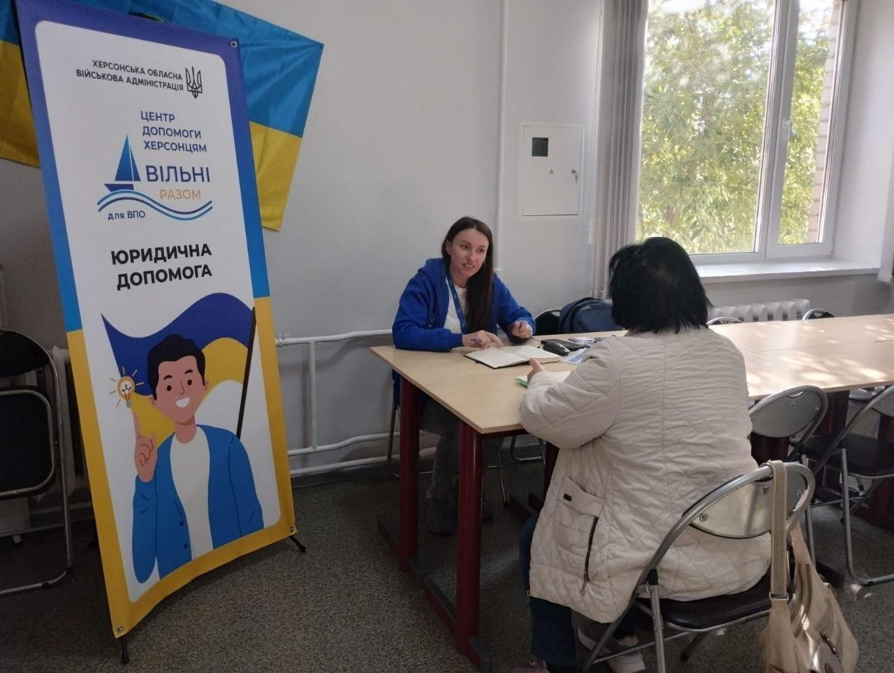 Юрисконсульт благодійного фонду «Право на захист» проводить юридичну консультацію для відвідувачки в житомирському координаційному центрі «Вільні разом», який системно підтримує переселенців з Херсонщини. У світлому кабінеті за дерев'яним столом сидить урядова фахівчиня у синьому худі з блокнотом, яка спілкується з жінкою-переселенкою у світлій куртці. Ліворуч у кімнаті встановлено великий фірмовий банер ХОВА з написом «ЮРИДИЧНА ДОПОМОГА», над яким закріплено Державний прапор України, а праворуч помітно велике вікно з видом на дерева. Під час цієї правової зустрічі внутрішньо переміщені особи отримали індивідуальні юридичні консультації з актуальних питань, що допомагає херсонцям у вирішенні правових проблем, відновленні втрачених документів та захисті своїх прав в евакуації.