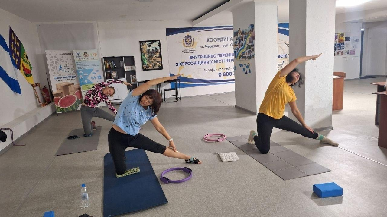 Три переселенки з Херсонщини зосереджено виконують вправи на гнучкість, займаючись стретчингом та йогою на фітнес-килимках у залі черкаського координаційного центру «Вільні разом». Жінки у зручному спортивному одязі роблять глибокий нахил тулуба вбік, використовуючи спеціальні пілатес-кільця та йога-блоки на підлозі. Заняття проходить у просторій світлій кімнаті хабу на тлі великого офіційного банера для внутрішньо переміщених херсонців у місті Черкаси та розвішаних Державних прапорів поруч із банером із зображенням кавуна. Ця оздоровча ініціатива, яка також включає заняття із зумби, організована для системної підтримки, покращення фізичного тонусу та емоційного розвантаження херсонських родин в евакуації.