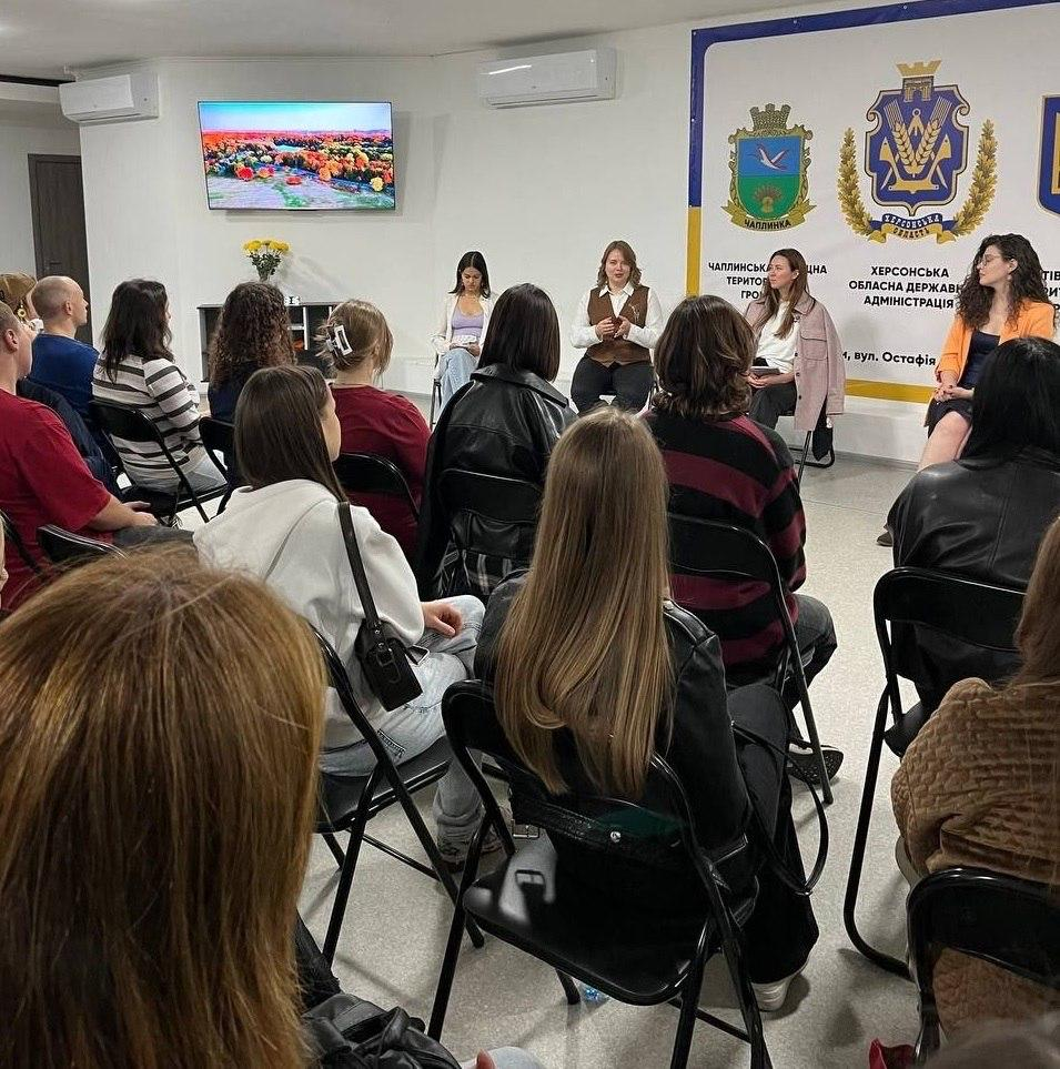 Переселенці з Херсонщини беруть участь у панельній дискусії до Всесвітнього дня ментального здоров’я, слухаючи виступи фахівчинь у залі черкаського координаційного центру «Вільні разом». Слухачі різного віку розсілися на стільцях півколом спиною до об'єктива камери, а навпроти них біля стіни сидять чотири експертки Простору підтримки ментального здоров’я «Cozy Mental Place», серед яких жінка у коричневій жилетці та дівчина у яскравому помаранчевому піджаку. Зустріч проходить у світлій кімнаті хабу, де ліворуч на стіні під кондиціонером працює телевізор із яскравим зображенням осіннього парку, а праворуч розгорнуто великий офіційний білий банер із гербами та написами Чаплинської селищної територіальної громади й Херсонської обласної державної адміністрації. Під час цього корисного заходу учасники детально обговорили психологічну стійкість під час тривалого стресу, а також долучилися до практичних майстерок, де власноруч виготовили метафоричні карти й антистресові вироби для покращення свого психоемоційного стану в евакуації.
