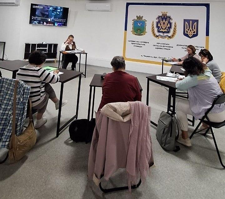 Переселенки з Херсонщини вдосконалюють мовні навички на занятті з англійської мови в черкаському координаційному центрі «Вільні разом», розсівшись за окремими темними столами у навчальній кімнаті. На передньому плані спиною до об'єктива кадру сидять учасниці у смугастій та бордовій кофтах, а праворуч інші відвідувачки роблять нотатки у робочих зошитах. Попереду за окремим столом під увімкненим настінним телевізором сидітиме викладачка у жилетці, яка координує навчальний процес, а на задній стіні кабінету розгорнуто великий офіційний білий банер із золотим Тризубом, гербами та назвами Чаплинської селищної громади, Долматівської сільської громади та Херсонської ОДА. Під час цього інтерактивного уроку учасниці занять говорили про живопис, скульптуру, музику, а також детально аналізували відому композицію Billy Joel «Piano Man», що допомогло херсонським родинам в евакуації суттєво розширити свій словниковий запас та вдосконалити розмовні навички в дружньому колі земляків.