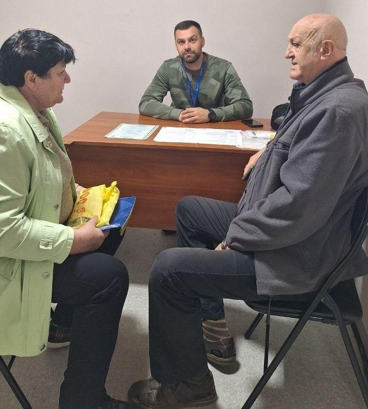 Фахівець громадської організації «Десяте квітня» проводить консультацію для переселенців із Херсонщини в черкаському координаційному центрі «Вільні разом». У кабінеті за дерев'яним столом сидить консультант у зеленому світшоті з синьою стрічкою на шиї, який розмовляє з відвідувачами старшого віку — жінкою у світло-зеленому пальті та чоловіком у сірій флісовій кофті. На столі перед фахівцем розкладено бланки заяв, офіційні папери у файлах та лежить телефон. Під час цієї правової зустрічі співробітник організації надав внутрішньо переміщеним особам роз'яснення щодо порядку подання заяв до Міжнародного реєстру збитків та допоміг підготувати необхідний пакет документів для захисту майнових прав херсонських родин в евакуації.