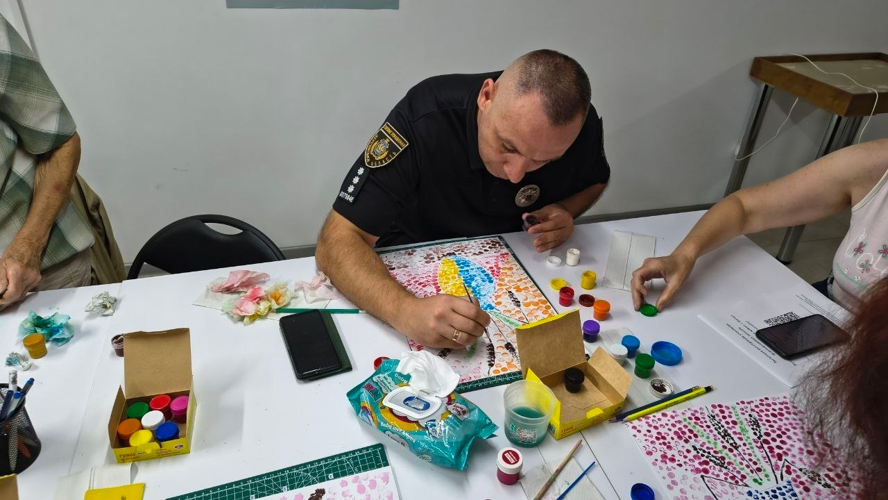 Співробітник поліції у чорній формі з офіційними шевронами та погонами капітана зосереджено малює разом з іншими учасниками за білим столом під час арттерапевтичного заняття «Натюрморт пальцями» в Центрі життєстійкості. Чоловік охайно наносить фарбу тонким пензликом на папір, розмальований яскравими крапками, ліворуч від нього за процесом стежить відвідувач у клітчастій сорочці, а праворуч жінка допомагає розставляти баночки з гуашшю. На робочій поверхні столу розкладено коробки з фарбами, пензлики, олівці, паперові серветки та мобільні телефони. Такі корисні заходи, які проводили в центрі протягом тижня, організовані для психоемоційного розвантаження, зняття внутрішньої напруги та покращення ментального здоров'я жителів громади.
