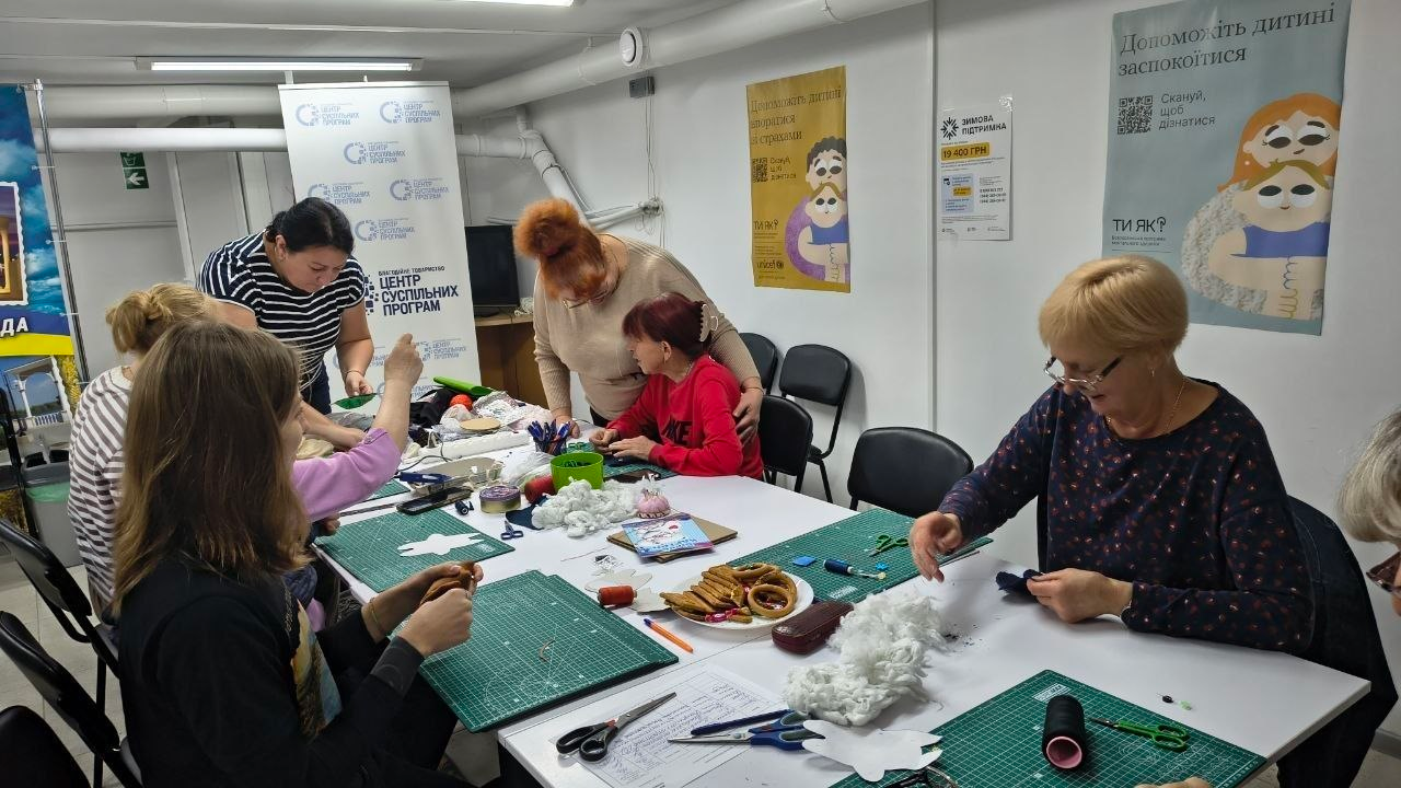 Учасниці артмайстерні «Киця» зосереджено працюють за великим столом під час арттерапевтичних заходів у Центрі життєстійкості, створюючи рукодільні м’які іграшки. Жінки різного віку розклали перед собою зелені макетні килимки й за допомогою ниток, ножиць та білого наповнювача зшивають фігурні тканинні заготівлі, а праворуч усміхнена жінка в окулярах охайно шиє виріб поруч із тарілкою з сушками. Заняття проходить у світлій кімнаті на тлі білого фірмовий банера БО «Центр суспільних програм» та інформаційних плакатів психологічної підтримки «Ти як?» на стіні. Проведені протягом тижня в центрі творчі зустрічі спрямовані на ефективне психоемоційне розвантаження, зняття внутрішньої напруги, розвиток дрібної моторики та всебічне покращення ментального здоров'я жителів громади.