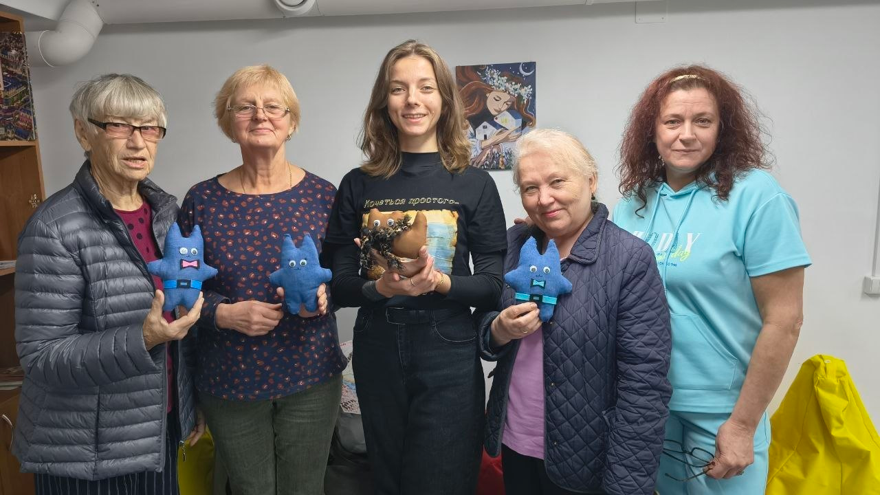 Учасниці артмайстерні «Киця» з усмішками позують для спільного фото, демонструючи виготовлені власноруч м’які іграшки після завершення арттерапевтичних заходів у Центрі життєстійкості. Жінки вишикувалися в один ряд перед об'єктивом камери та тримають у руках саморобних кумедних котів синього та коричневого кольорів з іграшковими пластиковими очима. По центру стоїть молода дівчина у чорному лонгсліві з малюнком, ліворуч від неї — двоє учасниць старшого віку в окулярах, а праворуч — жінка у синій стьобаній куртці та майстриня-організатор у яскраво-бірюзовому спортивному костюмі на тлі світлої стіни кімнати з малюнком. Проведені протягом тижня в центрі творчі зустрічі спрямовані на ефективне психоемоційне розвантаження, зняття внутрішньої напруги, розвиток дрібної моторики та всебічне покращення ментального здоров'я жителів громади.