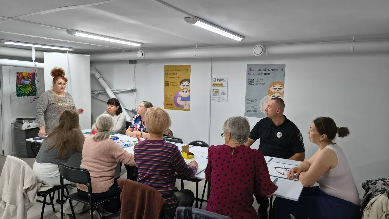 Учасники творчого заняття з малювання «Натюрморт пальцями» зібралися за великим білим столом у просторій кімнаті під час арттерапевтичних та психологічних заходів у Центрі життєстійкості. Ліворуч біля білої мобільної перегородки стоїть пишноволоса ведуча-майстриня в окулярах та сірій кофті, яка координує процес і розмовляє з присутніми жінками та співробітником поліції у чорній формі з офіційними шевронами Національної поліції України, які розсілися навколо робочих місць. На білій поверхні столу розкладено аркуші паперу, фарби та олівці, а на задній стіні кабінету під лінійними світильниками висять інформаційні плакати програми психологічної підтримки «Ти як?» та Дитячого фонду ООН (UNICEF). Проведені протягом тижня в центрі заходи спрямовані на ефективне психоемоційне розвантаження жителів громади, зняття внутрішньої напруги, розвиток творчих здібностей та всебічне зміцнення ментального здоров'я.