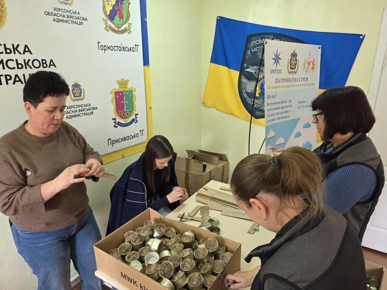 Волонтери та відвідувачі координаційного центру «Вільні разом» у Дніпрі, який надає всебічну підтримку переселенцям з Херсонщини, долучилися до допомоги українським захисникам, готуючи матеріали для окопних свічок. У кімнаті хабу за двома зсунутими столами працюють жінки: ліворуч відвідувачка у коричневому светрі та синіх джинсах розправляє картон, поруч дівчина у синій кофті скручує гофровані смужки, а на передньому плані інші учасниці вкладають заготовки в бляшанки біля великої картонної коробки, вщент заповненої підготовленими свічками. Робота проходить у патріотично оформленому кабінеті, де на стінах розвішано великий білий банер Херсонської ОВА з гербами Горностаївської та Присиваської територіальних громад, Державний прапор України, темний військовий стяг бригади та інформаційний плакат про патронатні сім'ї. Ця спільна благодійна ініціатива допомагає внутрішньо переміщеним херсонцям об'єднати зусилля в евакуації, підтримати ЗСУ та зробити свій важливий внесок у наближення перемоги.