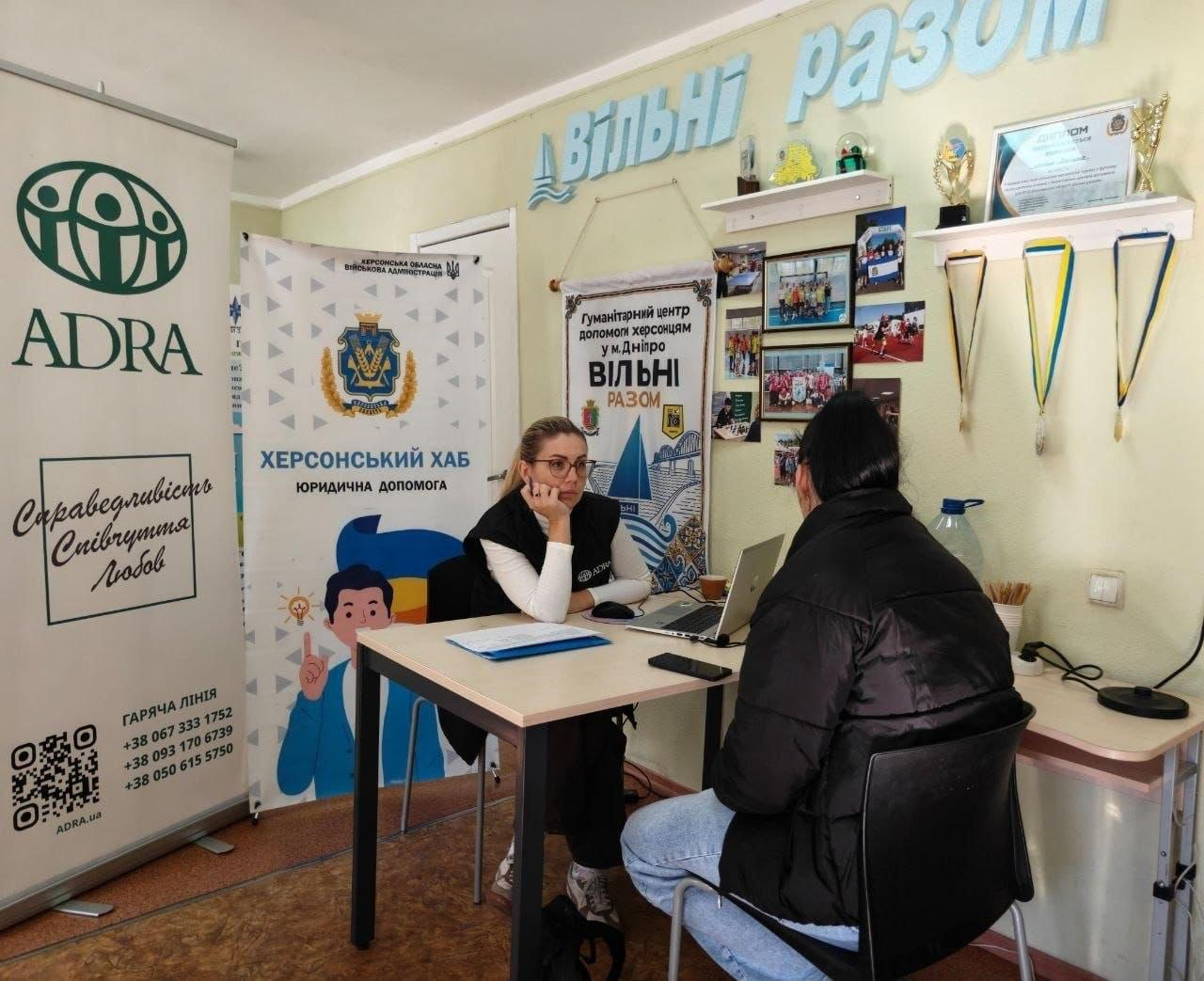 Координаційний центр «Вільні разом» у Дніпрі надає всебічну підтримку переселенцям з Херсонщини, організувавши прийом, під час якого юрисконсульти провідних благодійних фондів, зокрема «ADRA Ukraine» та «Право на захист», надали 19 індивідуальних консультацій. На світлині фахівчиня в окулярах та чорній жилетці з логотипом «ADRA» проводить зустріч із відвідувачкою в темній зимовій куртці за дерев'яним столом із ноутбуком. Ліворуч у кабінеті встановлено великий білий банер організації ADRA та фірмовий стенд ХОВА «ХЕРСОНСЬКИЙ ХАБ. ЮРИДИЧНА ДОПОМОГА», а на задній стіні під блакитним написом «Вільні разом» висить інформаційний плакат гуманітарного центру з фотоколажем та поличками з кубками й медалями. Така спільна робота правозахисників допомагає внутрішньо переміщеним херсонцям оперативно отримати кваліфіковану правову допомогу, розібратися з відновленням документів чи оформленням виплат і знайти дієву підтримку в евакуації.