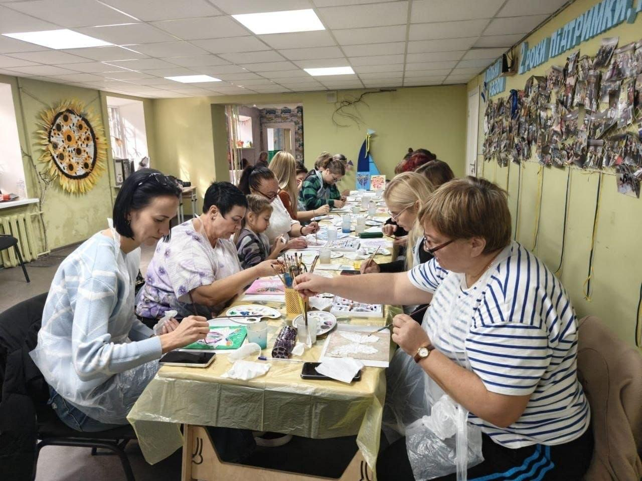 Переселенки з Херсонщини разом із маленькою дитиною зосереджено працюють над створенням об'ємних виробів під час арттерапевтичного заняття «Подих метелика» від ГО «Дівчата» у дніпровському координаційному центрі «Вільні разом». Жінки різного віку розсілися вздовж довгого спільного столу, застеленого захисною плівкою, та за допомогою пензликів, палітр і фарб охайно розмальовують фігурні 3D-картини у вигляді метеликів. Навчання проходить у світлій залі хабу з оливковими стінами, де ліворуч куток кімнати задекоровано великим соняшником, а на правій стіні під блакитним об'ємним написом розгорнуто великий панорамний фотоколаж зі світлинами з життя центру. Ця корисна творча ініціатива, організована дніпровським хабом для всебічної підтримки родин ВПО, поєднувала малювання з опануванням дієвих технік релаксації, що допомагає учасницям ефективно зняти внутрішню напругу, подолати стрес від евакуації та покращити ментальне здоров'я.