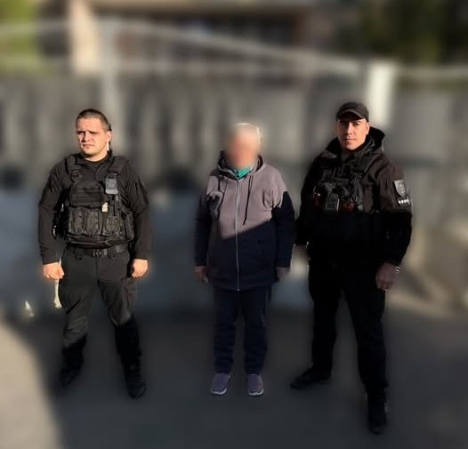 Двоє поліцейських офіцерів громади у чорній тактичній формі та бронежилетах позують для спільного фото на вулиці разом із евакуйованою цивільною особою з прифронтової Дар’ївської громади. По центру між правоохоронцями стоїть літня людина з розмитим обличчям у сірій кофті-худі, темних штанях та кросівках, а знімок зроблено вдень на асфальтованій дорозі на тлі приватного сектору з парканом. Поліцейські допомогли громадянину залишити небезпечну зону, яка постійно перебуває під загрозою ворожих обстрілів, та безпечно доставили його до транзитного центру у Херсоні. Там людина отримає необхідну допомогу, безпеку та подальше безкоштовне розміщення у безпечних регіонах, а для організації виїзду мешканці можуть цілодобово звертатися на «гарячу лінію» Херсонської ОВА.