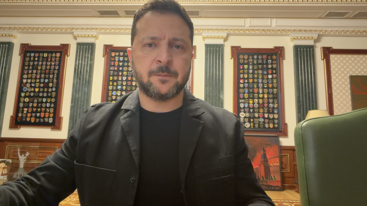 Президент України Володимир Зеленський у чорному піджаку та чорній футболці виступає з офіційним вечірнім зверненням до нації, перебуваючи у робочому кабінеті Офісу Президента. Голова держави зафіксований крупним планом по центру кадру на тлі білих стін із золотим ліпним карнизом, великих колон та трьох вертикальних стендів із військовими шевронами у дерев'яних рамках, а праворуч помітно спинку великого зеленого шкіряного крісла. У своєму виступі Володимир Зеленський анонсував нові пакети підтримки від Данії, Литви та Великої Британії, а також відкриття нових переговорних кластерів із Євросоюзом до кінця року. Президент прокоментував небезпечний удар п'ятьма балістичними ракетами по Києву, наголосивши на важливості оперативної розвідувальної взаємодії з партнерами, та критично оцінив незадовільний рівень підготовки столиці до зими й захисту інфраструктури, закликавши прискорити виконання планів стійкості за прикладом Харкова та Запоріжжя.