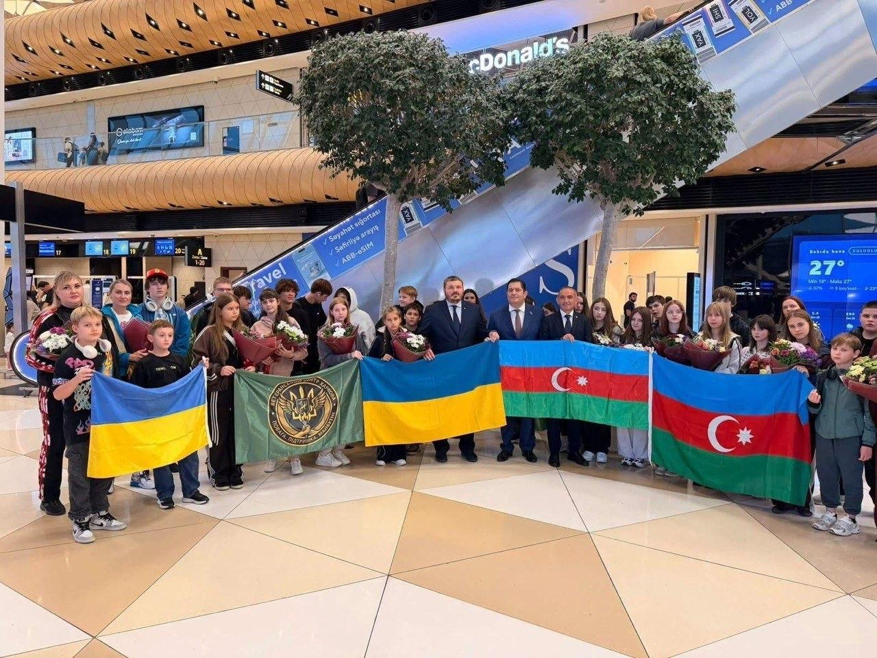 Велика група херсонських дітей з букетами квітів разом із представниками Посольства України та азербайджанськими партнерами позує для спільного фото в залі Міжнародного аеропорту в Баку, тримаючи розгорнуті національні прапори обох держав. Учасники зустрічі вишикувалися в довгий ряд під декоративними деревами, тримаючи перед собою два синьо-жовті прапори України, два синьо-червоно-зелені прапори Азербайджану та зелений стяг координаційного центру ХОВА з золотим Тризубом. Цю поїздку для дітей чинних та важкопоранених військовослужбовців організували управління у справах ветеранів та Департамент інфраструктури Херсонської ОДА на запрошення Посольства України. Під час відпочинку юні жителі Херсонщини пройшли курс медичної й психологічної реабілітації в Габалінському центрі, займалися спортом та відвідали численні історичні й розважальні локації, що сприяло їхньому емоційному відновленню.