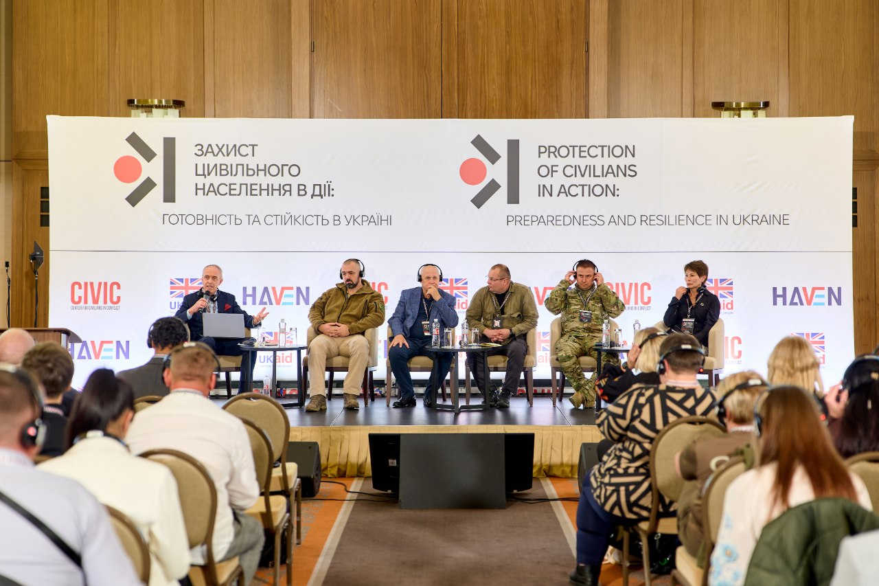 Група спікерів міжнародної конференції «Захист цивільного населення на практиці: готовність і стійкість в Україні» сидить на сцені в кріслах під час панельної дискусії в залі Києва. Експерти, серед яких модератор у костюмі з ноутбуком, фахівці в тактичному одязі, військовослужбовець ЗСУ в камуфляжі та жінка праворуч, використовують навушники синхронного перекладу, а перед ними розставлені столики з водою. Обговорення проходить на тлі великого білого банера з двомовними написами «ЗАХИСТ ЦИВІЛЬНОГО НАСЕЛЕННЯ В ДІЇ» та логотипами «CIVIC HAVEN» і «UK aid» на дерев'яній стіні. Херсонщину на цьому заході представила співробітниця ХОДА Ірина Осіпова, а ключовими темами наради громад, влади та сил безпеки стали евакуація, цивільно-військове співробітництво, адаптація систем оповіщення й організація захисту об'єктів критичної інфраструктури прифронтових територій в умовах атак безпілотників.