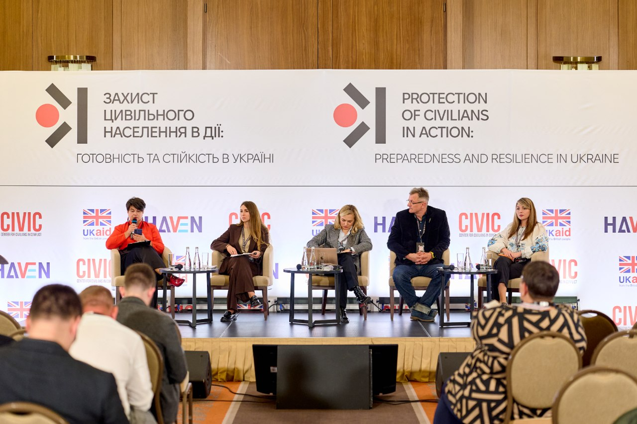 П'ять експертів міжнародної конференції з цивільного захисту міжвідомчих груп сидять на сцені під час панельної дискусії в залі Києва, де крайня праворуч — завідувачка сектору координації заходів у сфері захисту критичної інфраструктури Департаменту з питань цивільного захисту та оборонної роботи ХОДА Ірина Осіпова у білій вишиванці. Разом із представницею Херсонщини на підмостках перебувають ще три жінки та чоловік, які працюють із робочими документами й навушниками синхронного перекладу на тлі великого офіційного банера «ЗАХИСТ ЦИВІЛЬНОГО НАСЕЛЕННЯ В ДІЇ» з логотипами «CIVIC HAVEN» та «UK aid». Під час обговорення представники прифронтових громад, центральної влади та сил безпеки зосередилися на практичних рішеннях для територій, охопивши питання безпечної евакуації, цивільно-військового співробітництва, адаптації систем оповіщення й безпосередньої організації захисту об'єктів критичної інфраструктури в умовах постійних атак безпілотників.