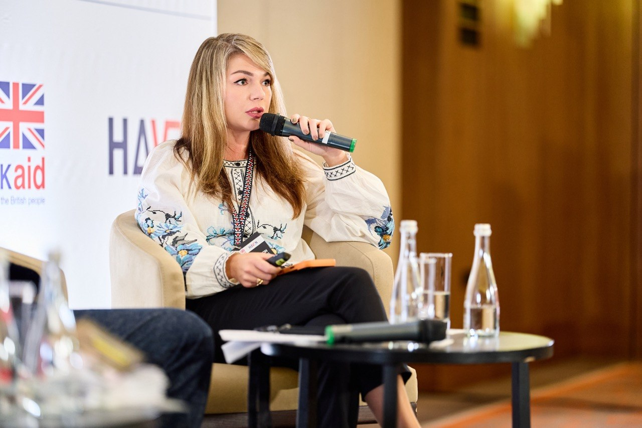 Завідувачка сектору координації заходів у сфері захисту критичної інфраструктури Департаменту з питань цивільного захисту ХОДА Ірина Осіпова виступає на сьомій міжнародній конференції «Захист цивільного населення на практиці: готовність і стійкість в Україні» у Києві. Посадовець сидить у м'якому кріслі, вбрана в білу вишиванку з блакитно-чорним орнаментом та чорні штани, і тримає в руці мікрофон, а на задньому плані помітно білий банер із логотипом «UK aid» на тлі дерев'яних панелей залу. Під час панельних дискусій представниця Херсонщини поділилася практичними рішеннями для захисту цивільних, обговоривши евакуацію, адаптацію систем оповіщення, ризики через активне застосування безпілотників та організацію роботи із захисту критичної інфраструктури в прифронтовому регіоні.