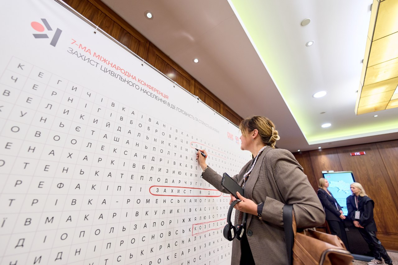 Учасниця конференції «Захист цивільного населення на практиці: готовність і стійкість в Україні» маркером обводить слова на інтерактивній стіні-кросворді з назвами українських громад. Жінка в коричневому піджаку з бейджем на шиї зафіксована у профіль біля білого панно, де червоним кольором уже виділено гасло «ЗАХИСТ В ДІЇ» та назви міст, зокрема Полтава, Ромни й Запоріжжя, а на задньому плані спілкуються дві дівчини біля цифрового екрана. Херсонщину на заході представила завідувачка сектору координації заходів у сфері захисту критичної інфраструктури Департаменту з питань цивільного захисту та оборонної роботи ХОДА Ірина Осіпова. Головними темами наради двохсот учасників у Києві стали евакуація цивільних, адаптація систем оповіщення, цивільно-військове співробітництво та посилення безпекових протоколів критичної інфраструктури в умовах постійних атак безпілотників.