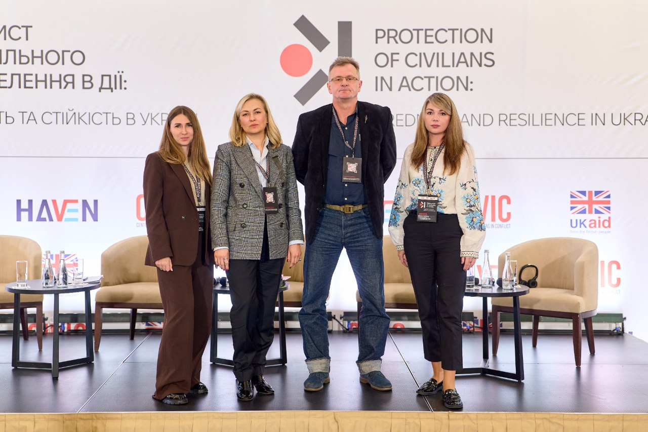 Чотири учасники міжнародної конференції з цивільного захисту з бейджами на шиї вишикувалися в ряд на сцені залу в Києві для спільного фото на повний зріст. По центру стоїть високий чоловік у темному піджаку та джинсах, ліворуч — дві жінки у коричневому й сірому костюмах, а праворуч позує завідувачка сектору координації заходів у сфері захисту критичної інфраструктури Департаменту з питань цивільного захисту та оборонної роботи ХОДА Ірина Осіпова в білій вишиванці з блакитним орнаментом. Знімок зроблено на тлі великого білого банера з написом «PROTECTION OF CIVILIANS IN ACTION» та логотипами «CIVIC HAVEN» і «UK aid», де позаду помітно бежеві крісла та журнальні столики. Цей захід об'єднав понад дві сотні представників громад, влади та сил безпеки для напрацювання практичних рішень щодо цивільно-військового співробітництва, евакуації, адаптації систем оповіщення та посилення протоколів захисту об'єктів критичної інфраструктури прифронтових територій в умовах атак безпілотників.
