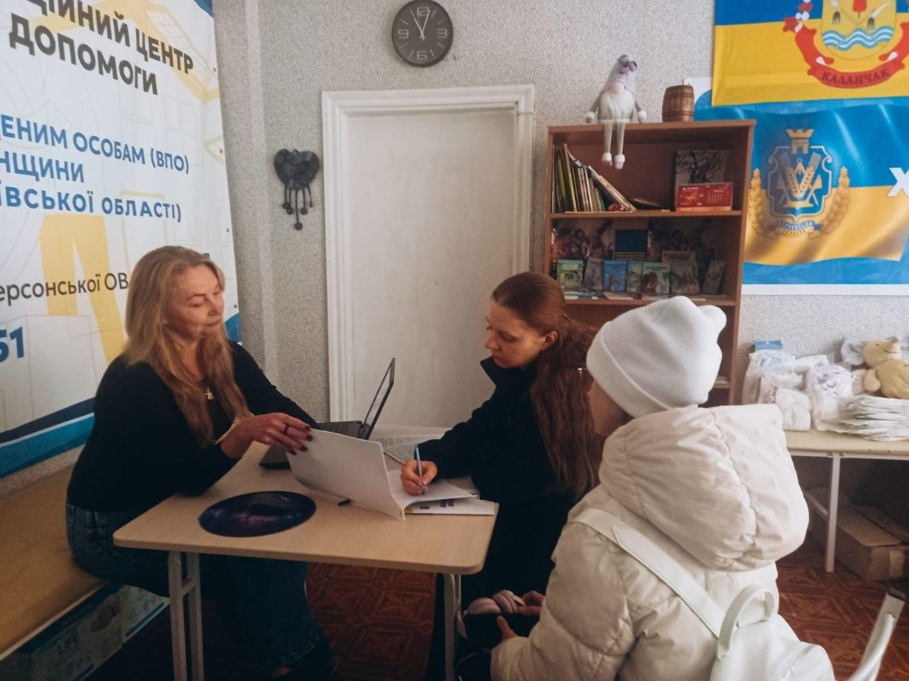 У київському координаційному центрі «Вільні разом» фахівці проводять індивідуальну консультацію з працевлаштування та юридично-соціального супроводу виплат для родини переселенців із Херсонщини. Ліворуч за світлим столом сидить консультант у чорній кофті поруч із відкритим ноутбуком і бланком документів, а рудоволоса фахівчиня робить нотатки в журналі. Навпроти них на стільці сидітиме дівчинка-відвідувачка у світлій зимовій куртці та білій шапці. Консультація проходить у кабінеті хабу, де ліворуч встановлено інформаційний банер ХОВА, на задній стіні помітно настінний годинник над білими дверима, а праворуч розташовано шафу з книгами та прапори України й Каланчацької громади. Ця комплексна консультативна допомога, організована для всебічної підтримки внутрішньо переміщених осіб, допомагає херсонським родинам в евакуації оперативно вирішувати питання життєзабезпечення, знаходити роботу та успішно оформлювати належні державні виплати.