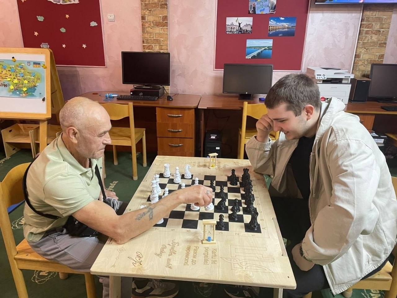 Ужгородський координаційний центр «Вільні разом» системно підтримує переселенців з Херсонщини, провівши засідання шахового клубу «Переможний шах» на тему «Логіка переможця». На світлині двоє чоловіків грають у шахи за світлим дерев'яним столом із вигравіруваним Тризубом і гаслом «Разом до перемоги!». Ліворуч сидить літній чоловік у світлій футболці, який робить хід білою фігурою, а навпроти нього праворуч розмістився усміхнений юнак у білій куртці, який грає чорними фігурами. Зустріч проходить у кабінеті хабу на тлі комп'ютерних столів, дитячої мапи України та бордових настінних стендів із фотографіями херсонських краєвидів. Така інтелектуальна ініціатива допомагає внутрішньо переміщеним херсонцям різного віку цікаво проводити дозвілля, спілкуватися з земляками та отримувати необхідне психоемоційне розвантаження в евакуації.
