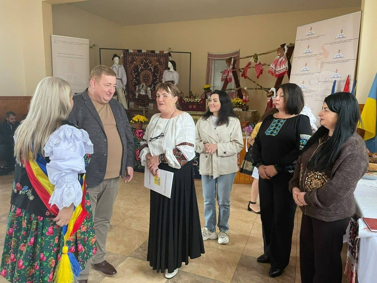 Керівниця ужгородського координаційного центру «Вільні разом» долучилася до міжнародного фестивалю «Міжетнічне співіснування в Сату-Маре — традиційне українське кулінарне мистецтво», що об'єднав понад 150 учасників з Румунії та України. На світлині зафіксовано спілкування делегацій, де по центру стоїть керівниця ужгородського хабу в білій вишиванці та довгій чорній спідниці, а на її грудях на мотузці висять чорні окуляри. Вона тримає інформаційну папку і розмовляє з чоловіком у сірому піджаку та румунською представницею в етнічному вбранні на тлі автентичних килимів, рушників і манекенів у народному одязі. Програма заходу в повіті Сату-Маре включала дегустацію традиційних українських страв, музичні виступи та культурний обмін, спрямований на зміцнення міжнаціональних зв'язків та всебічну підтримку переселенців.