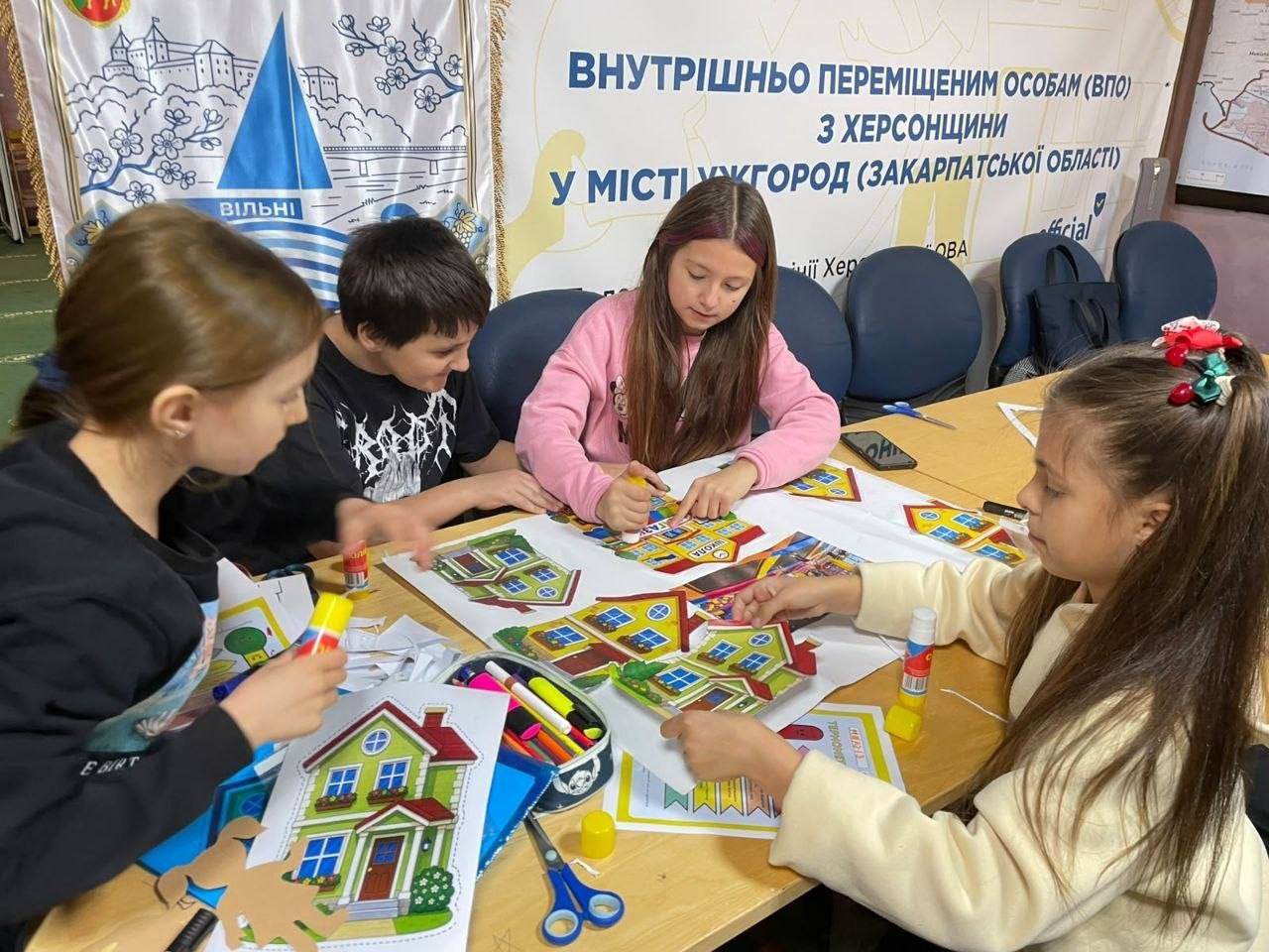 Ужгородський координаційний центр «Вільні разом» системно підтримує переселенців з Херсонщини, організувавши для 6 юних учасників зустріч групи психологічної підтримки «Створюємо своє місто», яку провела фахівчиня ГМ «Проліска». На світлині діти-переселенці у повсякденному одязі зосереджено створюють паперові аплікації у вигляді затишних кольорових будиночків навколо дерев'яного столу з фломастерами, клеєм і ножицями на тлі офіційного банера хабу та прапора «Вільні разом». Така корисна психосоціальна ініціатива допомагає малечі в евакуації емоційно розвантажитися, подолати стрес і висловити мрії про рідну домівку.