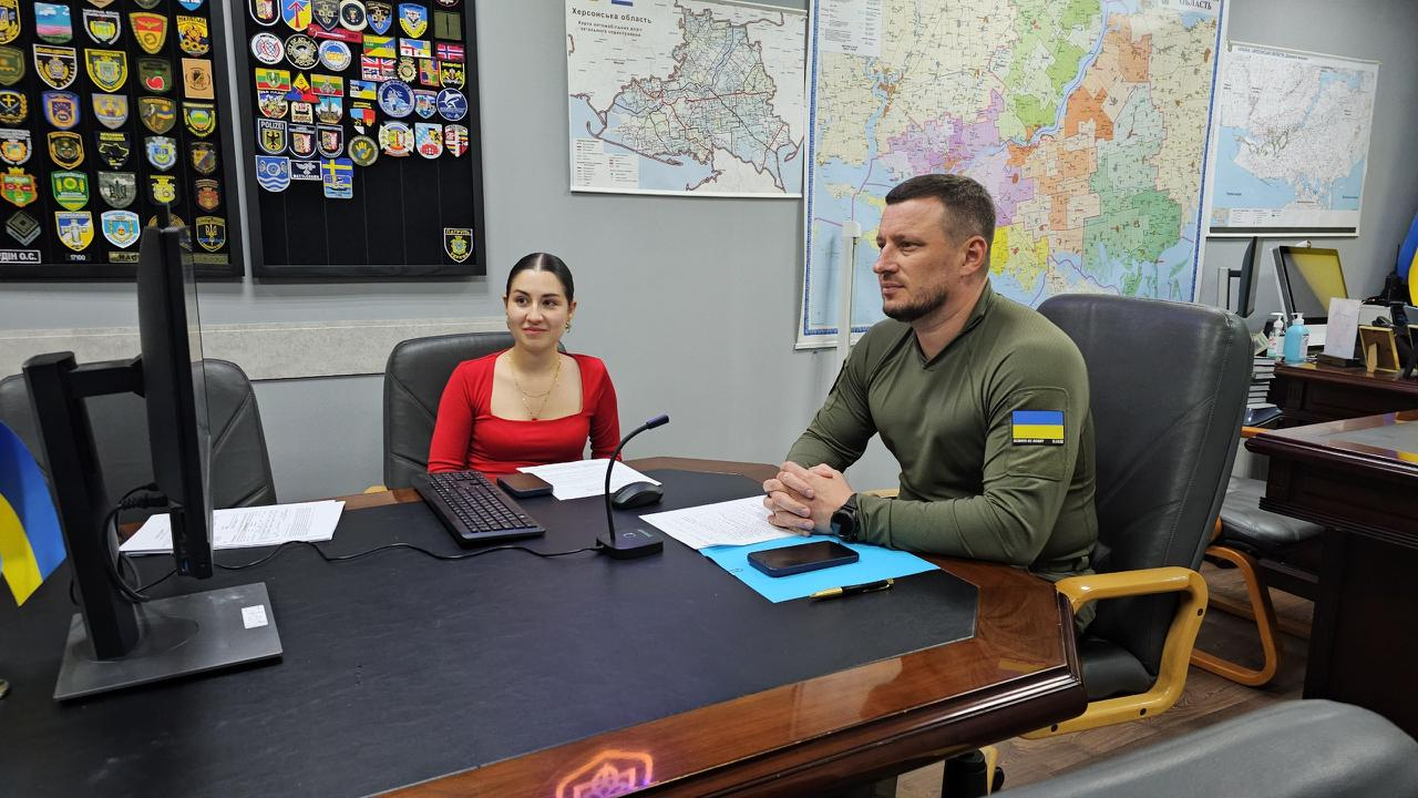 На світлині зафіксовано участь начальника Херсонської ОВА Олександра Прокудіна та начальниці управління міжнародного співробітництва Марії Богодухової в онлайн-зустрічі робочої групи Unbreakable Europe в Європейському парламенті. Вони сидять за великим дерев'яним столом і дивляться на монітор комп'ютера ліворуч. Олександр Прокудін одягнений у кофту кольору хакі з прапором України на плечі. На столі перед ними лежать документи, телефони, клавіатура, настільний мікрофон та встановлено маленький український прапорець. На сірих стінах кабінету позаду них розміщені карти, зокрема велика мапа Херсонської області, та великий стенд із військовими шевронами.