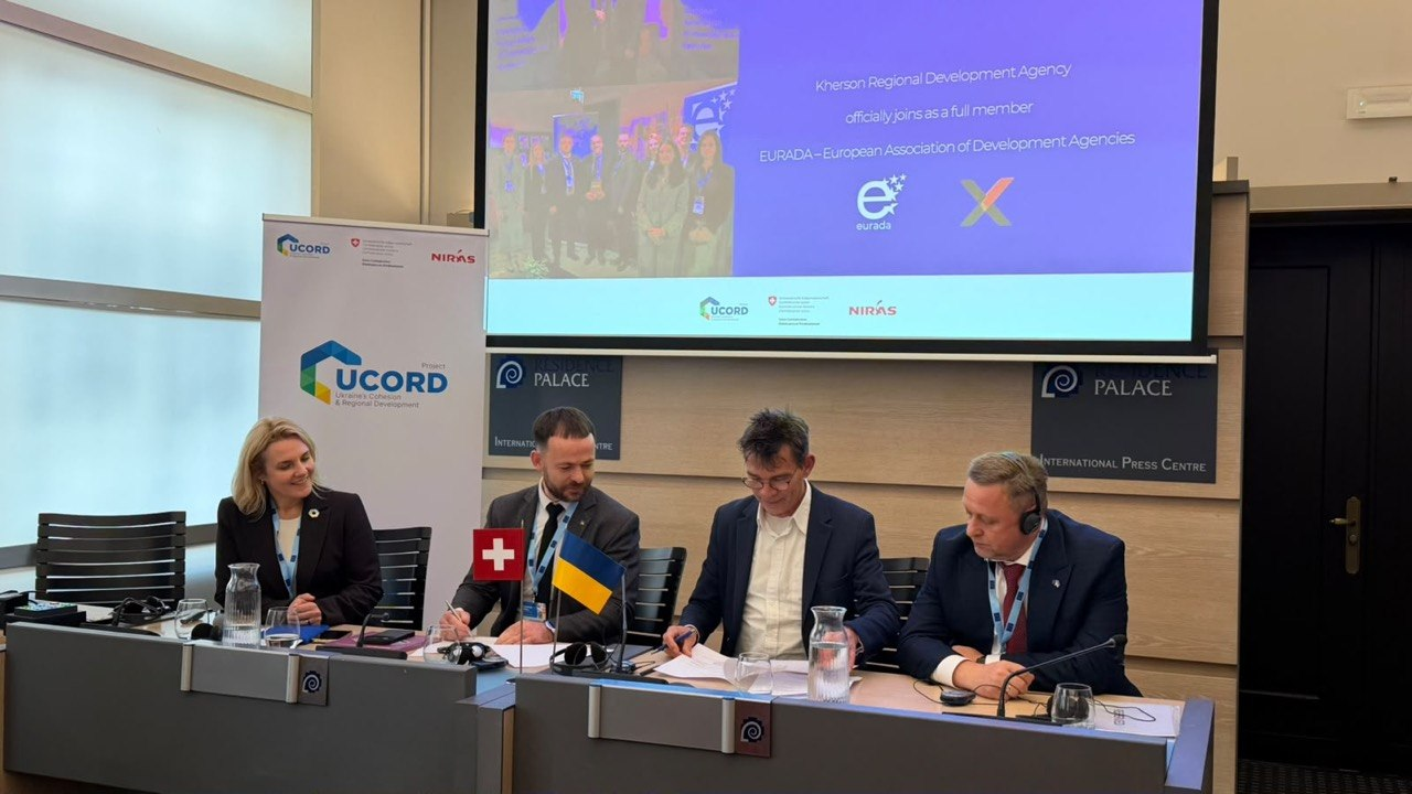 Херсонщина розширює співпрацю з європейськими інституціями під час European Week of Regions and Cities 2026 у Брюсселі, де Агенція регіонального розвитку «Офіс євроінтеграції» області офіційно увійшла до складу Європейської асоціації агенцій розвитку (EURADA). На столі пресцентру встановлено маленькі прапори Швейцарії й України та банер проєкту UCORD, а за процесом підписання на тлі великого мультимедійного екрана з інформацією про Херсонську агенцію спостерігають іноземні партнери та українська делегація. Ліворуч за сірим столом сидить представниця міжнародної організації у чорному жакеті, поруч із якою директор установи Олег Кулакевич робить нотатки ручкою, по центру європейський посадовець в окулярах завіряє офіційні папери, а праворуч перший заступник начальника Херсонської ОВА Дмитро Бутрій вивчає умови угоди. Ця інтеграція, профінансована проєктом UCORD за підтримки уряду Швейцарії, відкриває громадам прямий доступ до співпраці з регіонами країн ЄС для залучення фінансування та відбудови прифронтових територій.