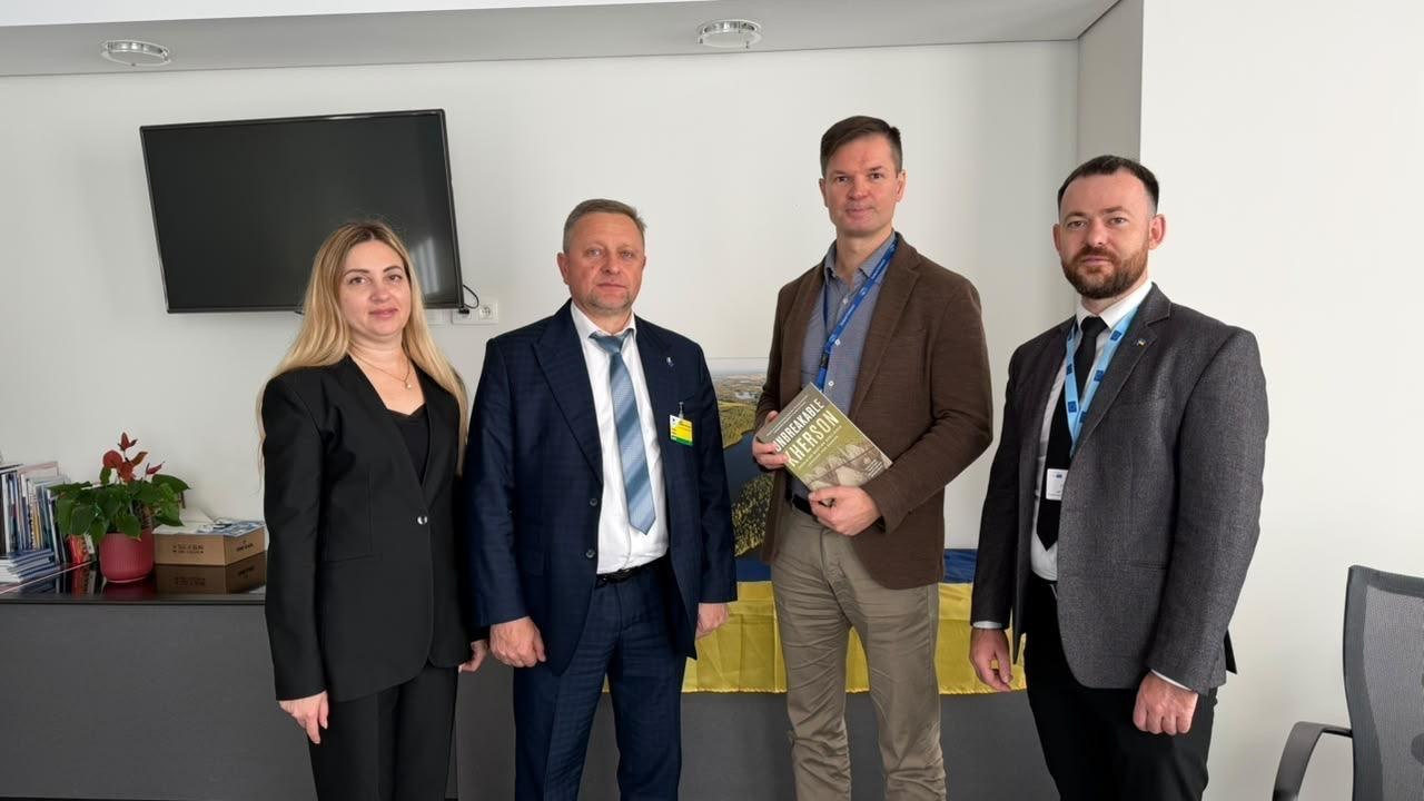 На світлині у світлому кабінеті зображено делегацію Херсонської області разом із європейським партнером під час Європейського тижня регіонів і міст у Брюсселі. У лінійку зліва направо стоять чотири особи: заступниця директора Агенції регіонального розвитку Інна Бутрій у чорному діловому костюмі, перший заступник начальника Херсонської ОВА Дмитро Бутрій у темно-синьому костюмі з краваткою та бейджем, депутат Європарламенту від Литви у коричневому піджаку, світлих штанях та з блакитною стрічкою-бейжем на шиї, а також директор Агенції Олег Кулакевич у сірому піджаку, чорній краватці та з бейджем. Литовський євродепутат тримає в руках та демонструє англомовну книгу-презентацію з написом «UNBREAKABLE KHERSON» на обкладинці. На задньому плані кадру на білій стіні висить вимкнений плазмовий телевізор, ліворуч на тумбі стоїть вазон із червоною квіткою та складені папери, а по центру за спинами присутніх видно Державний Прапор України.