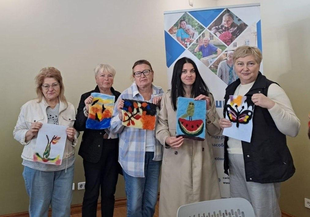 На світлині зображено п'ятьох жінок із числа внутрішньо переміщених осіб з Херсонщини, які тримають у руках свої роботи після майстер-класу з «Тканинної мозаїки» у Львівському координаційному центрі «Вільні разом». Учасниці заходу вишикувалися в один ряд у світлій кімнаті на тлі офісного інформаційного банера з фотографіями. Якщо дивитися від правого краю до лівого, перша жінка з короткою русявою зачіскою у чорному жилеті тримає мозаїку із зображенням метелика. Наступна учасниця з довгим темним волоссям у бежевому тренчі демонструє роботу із совою на шматочку кавуна. Третя жінка в окулярах та кардигані у клітинку тримає абстрактний кольоровий візерунок, а четверта учасниця у чорному піджаку показує пейзаж із заходом сонця та птахами. П'ята жінка ліворуч у світлій куртці тримає малюнок із рослинними мотивами на білому аркуші.