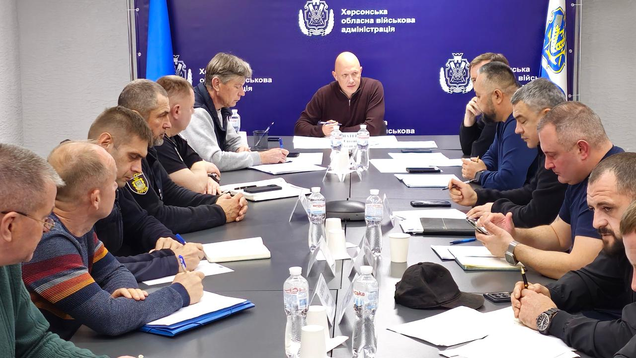 На світлині зображено хід робочої наради за участю заступника начальника Херсонської ОВА Євгенія Ігнатенка та представників відповідальних служб. Учасники зустрічі сидять один навпроти одного за довгим сірим столом у залі засідань. По центру на чолі столу сидить заступник начальника Херсонської ОВА Євгеній Ігнатенко в коричневій кофті, який сперся руками на стіл. Поруч із ним з одного боку столу сидить заступник міського голови Херсона Сергій Орєхов, одягнений у біло-синю кофту, а з іншого боку (де його майже не помітно за сусідами по столу) поруч сидить директор Департаменту з питань цивільного захисту та оборонної роботи Херсонської ОДА Євген Пісоцький. На столі перед усіма присутніми розкладено робочі папери, теки з документами, мобільні телефони, канцелярію, паперові стаканчики та пластикові пляшки з водою. За спиною головуючого розташований великий темно-синій брендований банер із повторюваним написом «Херсонська обласна військова адміністрація» та офіційним золотисто-блакитним гербом області, а ліворуч і праворуч від банера видно вертикально встановлені Державні Прапори України.