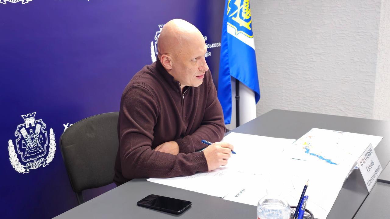 На світлині крупним планом зображено заступника начальника Херсонської ОВА Євгенія Ігнатенка під час проведення робочої наради щодо відновлення антидронових сіток на дорогах області. Посадовець сидить у профілі на сірому офісному кріслі за великим сірим столом і зосереджено дивиться вперед у бік учасників засідання. Він одягнений у коричневу кофту на блискавці та тримає у правій руці синю кулькову ручку. Перед ним на столі розкладено робочі документи, папери, лежить мобільний телефон, стоїть пластикова пляшка з водою та підставка з канцелярією. Також на робочій поверхні столу з правого боку розміщено схематичну географічну карту місцевості з позначеними лініями. По ліву руку від посадовця, праворуч у кадрі, видно частину вертикально встановленого прапора Херсонської області, а за його спиною розташований великий темно-синій брендований банер із золотисто-білим гербом.