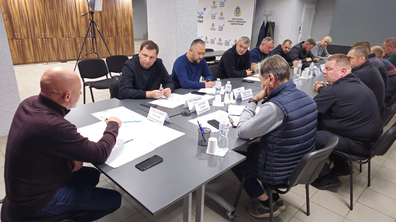 На світлині з ширшого ракурсу зображено хід робочої наради під керівництвом заступника начальника Херсонської ОВА Євгенія Ігнатенка. На передньому плані ліворуч, спиною до об'єктиву, сидить заступник начальника ОВА Євгеній Ігнатенко в коричневій кофті, який веде засідання. Безпосередньо поруч із ним по ліву руку за цією ж стороною столу сидить директор Департаменту з питань цивільного захисту та оборонної роботи ХОДА Євген Пісоцький у чорному світшоті. Навпроти нього, по іншу сторону столу (по праву руку від головуючого), сидить заступник міського голови Херсона Сергій Орєхов, одягнений у білий светр на блискавці та темно-синю жилетку поверх нього. На столі перед ними розкладено великі схематичні карти місцевості, папери, теки з документами, пластикові пляшки з водою та паперові стаканчики. На задньому плані зали засідань видно світло-сірі стіни, білий банер із логотипами «Official Media Center Херсонська обласна військова адміністрація», дерев'яні шафи ліворуч.