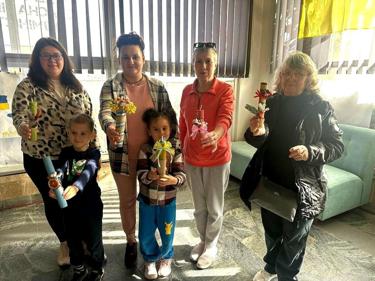 На світлині зображено чотирьох жінок та двох дітей із числа переселенців з Херсонщини, які тримають у руках власноруч виготовлені свічки з натуральної вощини у Миколаївському координаційному центрі «Вільні разом». Учасники заходу вишикувалися в один ряд у світлій кімнаті: якщо дивитися від правого краю до лівого, перша жінка в окулярах та чорній куртці тримає жовту свічку, наступна учасниця у кораловій кофті — червону свічку з бантиком, далі стоїть маленька дівчинка в синіх штанях із мамою, а ліворуч — маленький хлопчик із блакитною свічкою та жінка у плямистій кофті. Усі свічки мають унікальний декор із сухоцвітів, стрічок та ялинових гілочок. На задньому плані кадру видно великі вікна з вертикальними жалюзі, м'який світло-бірюзовий диван та елемент синьо-жовтого прапора.
