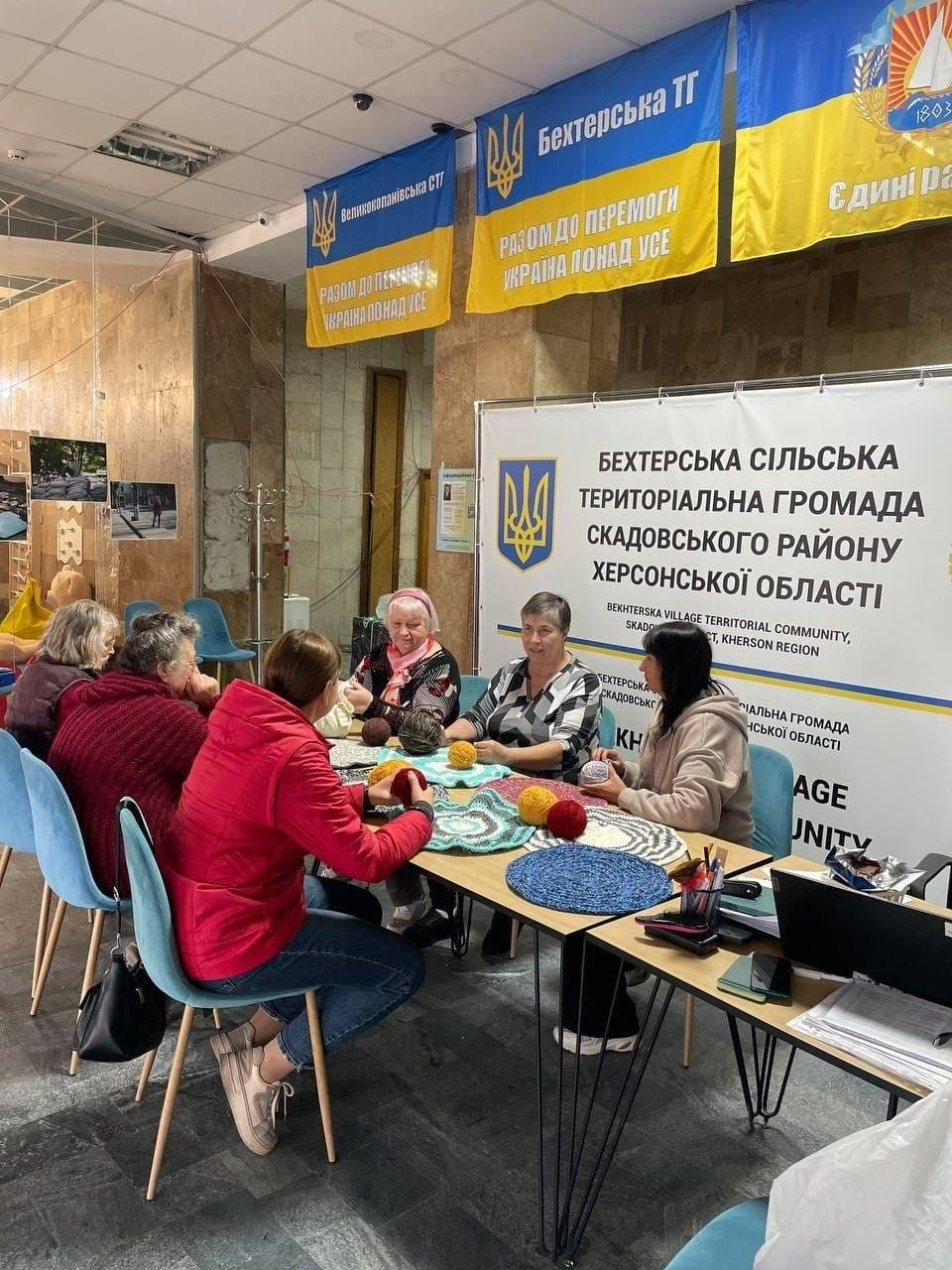 На світлині зображено жінок старшого та середнього віку, які сидять за великим столом у Миколаївському координаційному центрі «Вільні разом» під час занять у гуртку «Коло тепла». Учасниці зосереджено в'яжуть спицями та гачками круглі різнокольорові патріотичні килимки із товстої пряжі. На передньому плані ліворуч сидить жінка в яскраво-червоній куртці та синіх джинсах, яка змотує клубок жовто-гарячих ниток. Поруч із нею за столом розмістилися інші майстрині у бордовому светрі, фіолетовій куртці та чорно-білій блузці. На столі перед ними розкладено готові плетені вироби блакитного, синього та білого кольорів, лежать різнокольорові клубки вовни, спиці, ножиці та відкритий ноутбук праворуч. Робоче місце облаштоване на тлі офіційного білого стенда з написом «БЕХТЕРСЬКА СІЛЬСЬКА ТЕРИТОРІАЛЬНА ГРОМАДА СКАДОВСЬКОГО РАЙОНУ ХЕРСОНСЬКОЇ ОБЛАСТІ». Над столом під стелею підвішені великі синьо-жовті прапори Великокопанівської СТГ та Бехтерської ТГ з патріотичними гаслами «РАЗОМ ДО ПЕРЕМОГИ» та «УКРАЇНА ПОНАД УСЕ».