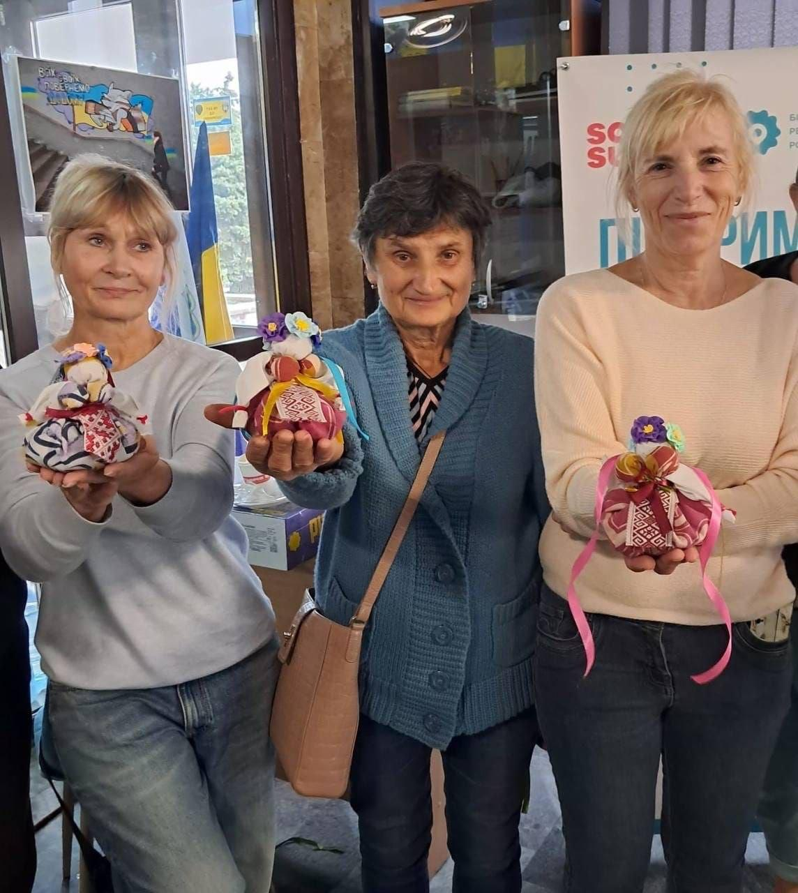 На світлині зображено жінок із числа внутрішньо переміщених осіб з Херсонщини, які тримають у руках власноруч виготовлені ляльки-травниці після майстер-класу від громадської організації «Білозерський центр регіонального розвитку». Учасниці творчого заняття стоять поруч у приміщенні Миколаївського координаційного центру «Вільні разом». Якщо дивитися від правого краю до лівого, перша жінка зі світлим волоссям у бежевому светрі та темних джинсах тримає на долоні ляльку з бордовою спідницею та рожевими стрічками. Поруч із нею стоїть жінка у синьому в'язаному кардигані на ґудзиках із коричневою сумкою через плече, яка демонструє оберегову ляльку з вишитими елементами та блакитними стрічками. Третя учасниця ліворуч у світло-сірому світшоті та блакитних джинсах показує свою травницю в пишній спідниці з орнаментом. На задньому плані кадру видно велике вікно, елемент банера з написами, а також патріотичний малюнок і Державний Прапор України.