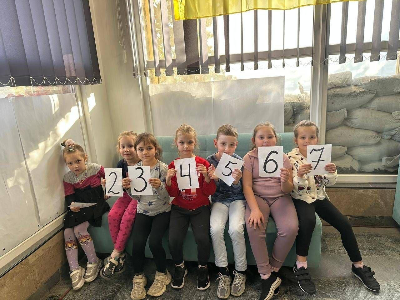 На світлині зображено дітей переселенців із Херсонщини під час заняття з підготовки до школи у Миколаївському координаційному центрі «Вільні разом». Вони сидять в один ряд на довгому бірюзовому дивані та тримають перед собою білі аркуші з великими чорними цифрами від «2» до «7». Хлопчики та дівчатка одягнені у повсякденний спортивний одяг і кофти різних кольорів. Простір освітлюється через великі вікна з вертикальними жалюзі, за якими на підвіконні з боку вулиці складено захисні мішки з піском.
