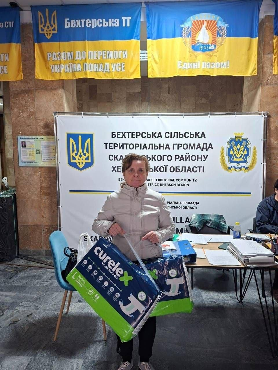 На світлині зафіксовано жінку з числа внутрішньо переміщених осіб з Херсонщини, яка отримує допомогу у Миколаївському координаційному центрі «Вільні разом». Вона стоїть по центру світлої зали в куртці світло-бежевого кольору та темних штанях і тримає велику упаковку засобів гігієни бренду curelex темно-синього кольору із зеленими елементами. Позаду неї встановлено великий білий офіційний стенд із написами «БЕХТЕРСЬКА СІЛЬСЬКА ТЕРИТОРІАЛЬНА ГРОМАДА СКАДОВСЬКОГО РАЙОНУ ХЕРСОНСЬКОЇ ОБЛАСТІ» та Державним Гербом України ліворуч. У верхній частині кадру під стелею підвішені великі синьо-жовті прапори Бехтерської територіальної громади та Лазурненської громади із гаслами «РАЗОМ ДО ПЕРЕМОГИ УКРАЇНА ПОНАД УСЕ» та «Єдині разом!». Праворуч на задньому плані помітно край робочого столу, на якому лежать теки з документами, папери та канцелярія, а також видно частину фігури працівника центру в темному одязі.